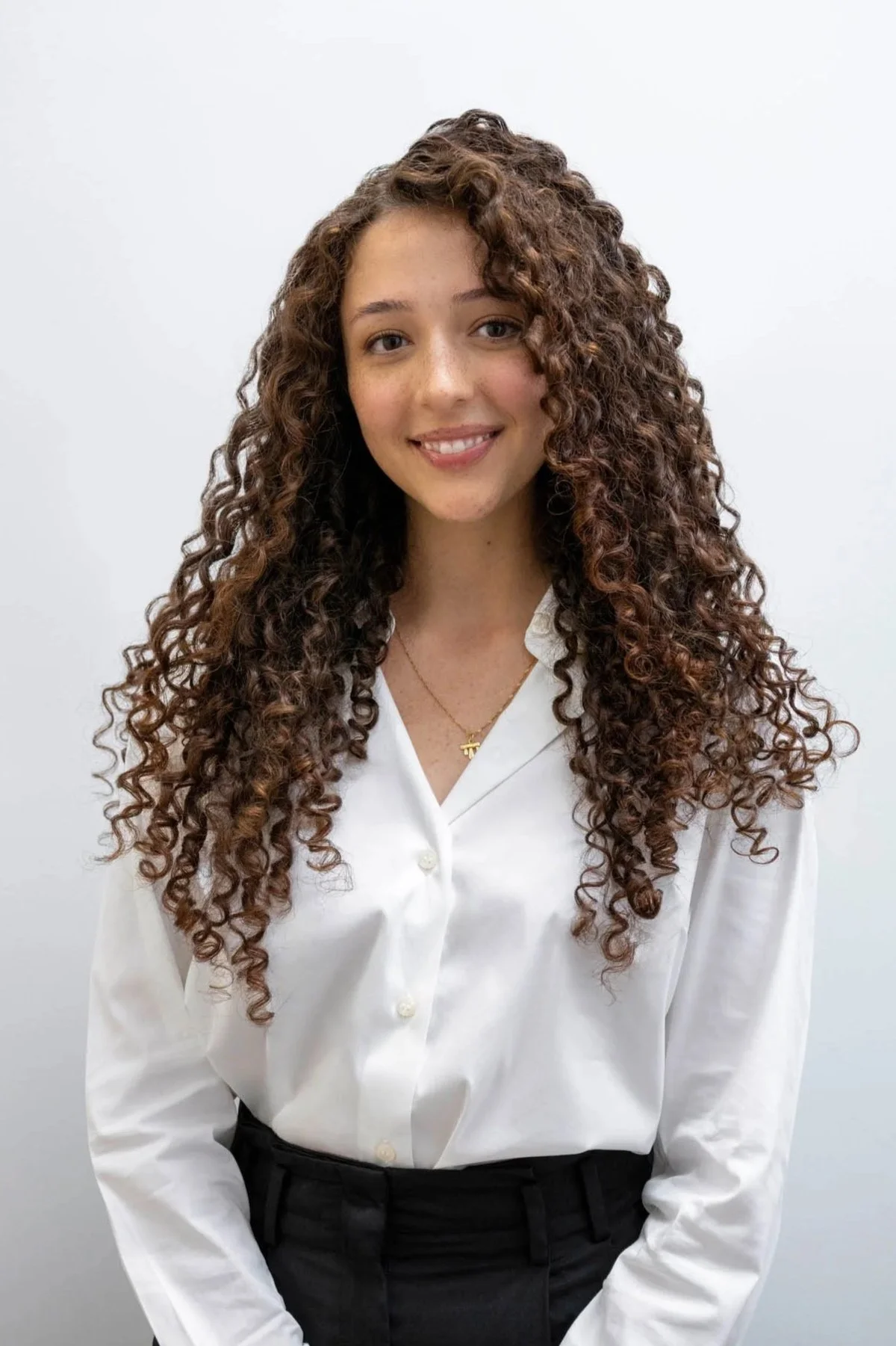 A young woman with long, curly brown hair smiling at the camera, wearing a white button-up shirt and black pants, standing against a plain white wall.