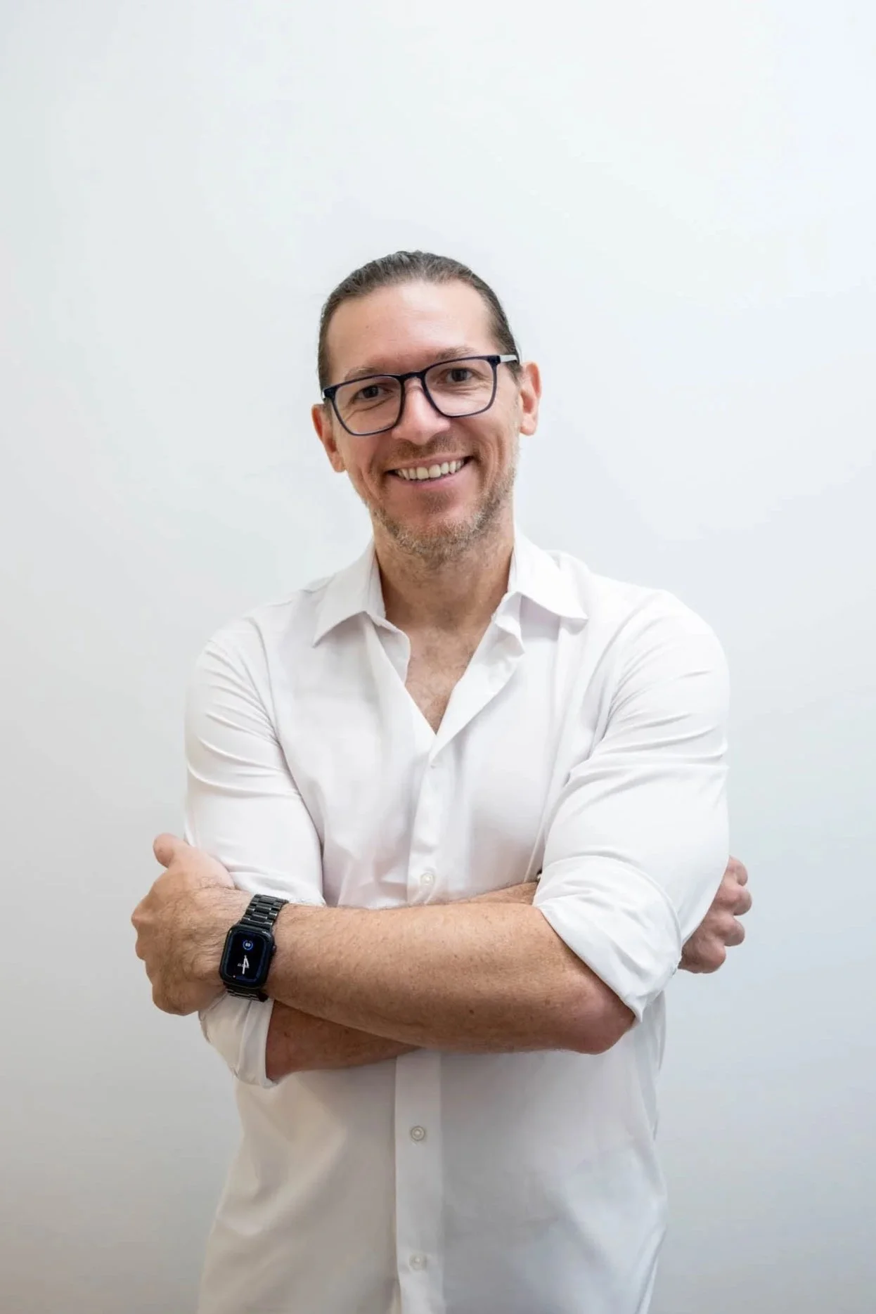 Smiling man with glasses and a beard wearing a white shirt and a smartwatch, standing with arms crossed against a plain white background.
