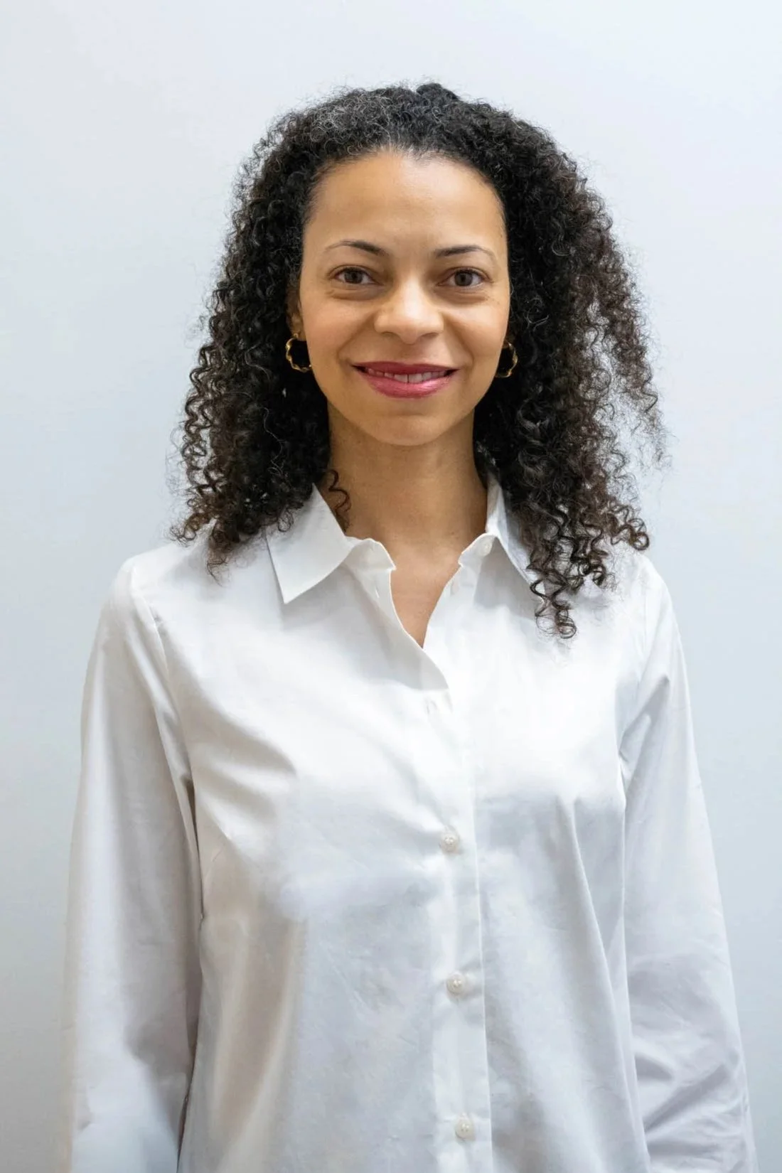 A woman with curly dark hair, wearing gold hoop earrings and a white button-up shirt, standing against a plain light-colored wall, smiling at the camera.