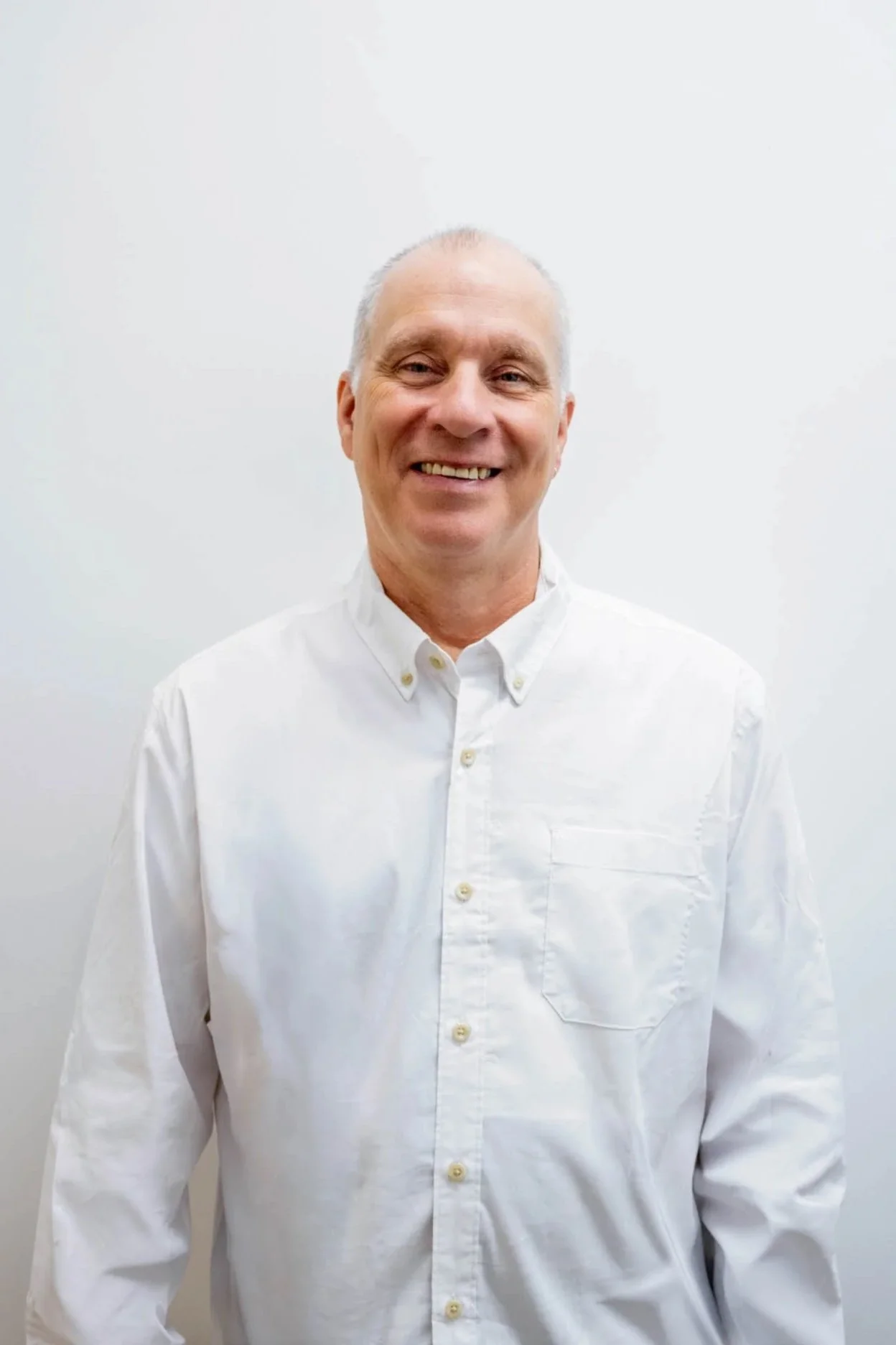 A smiling middle-aged man with short gray hair wearing a white button-up shirt standing against a plain white wall.