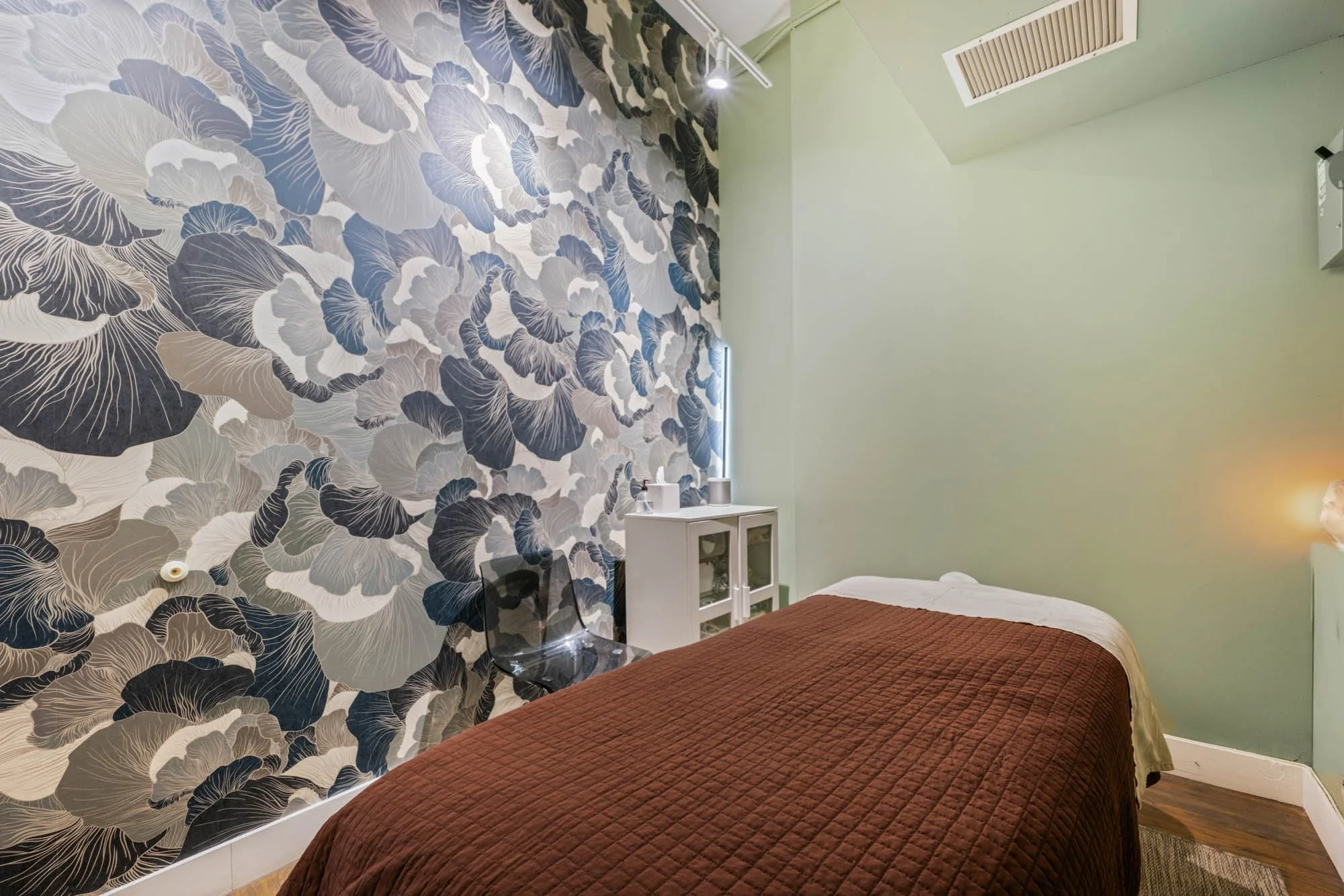 A bedroom with a floral accent wall, a single bed with a brown quilt, a transparent chair, and a small white cabinet. The room has a ceiling vent and dim lighting.