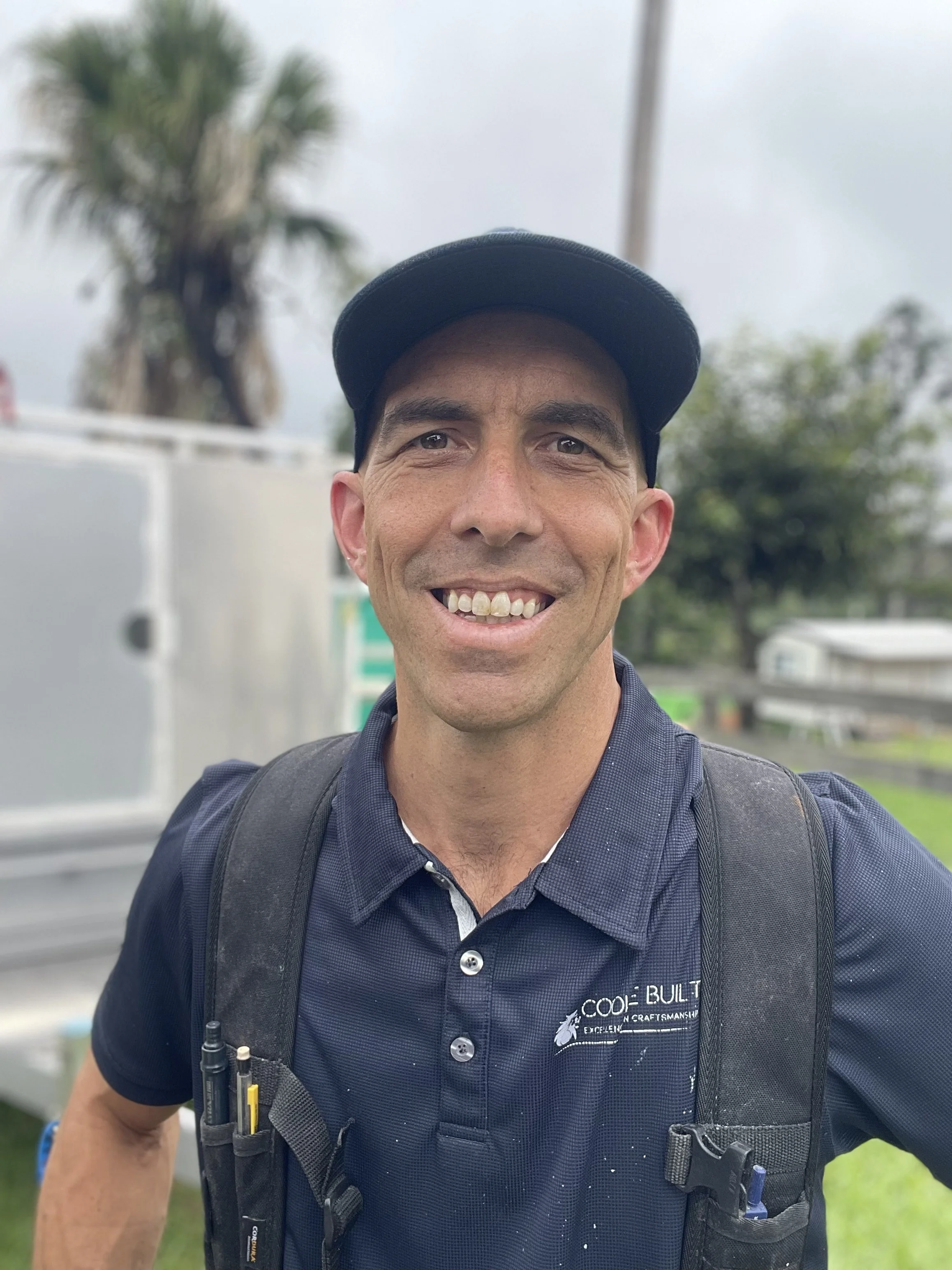 A smiling man wearing a black cap, navy polo shirt, and backpack outdoors with trees and a cloudy sky in the background.