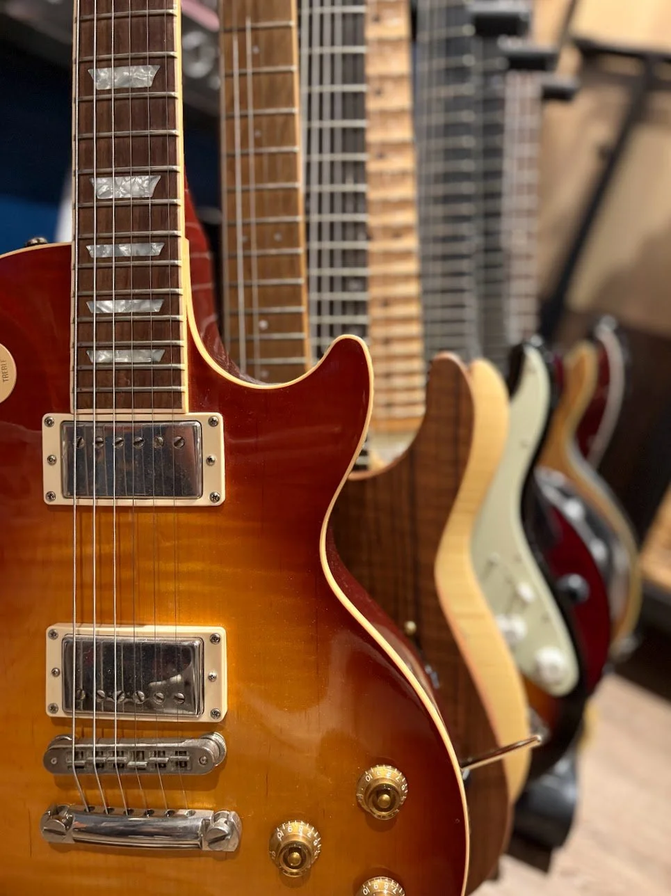 Close-up view of a row of electric guitars hanging on a wall, with focus on a sunburst guitar in the front. The guitars are in various colors and styles, with some having visible knobs and pickups.