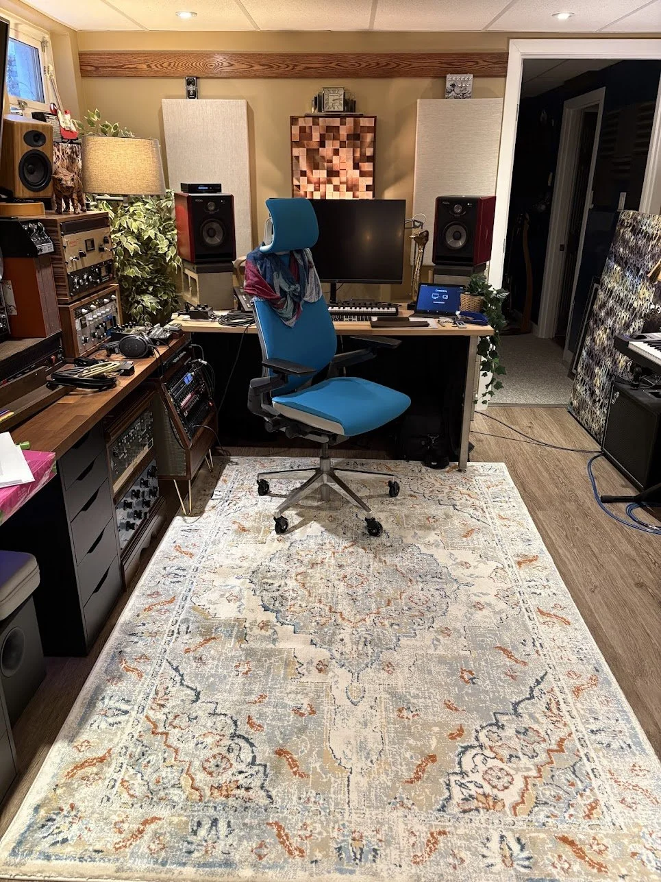 Home recording studio with a blue office chair, computer monitor, multiple audio speakers, sound equipment, and acoustic panels on the walls.
