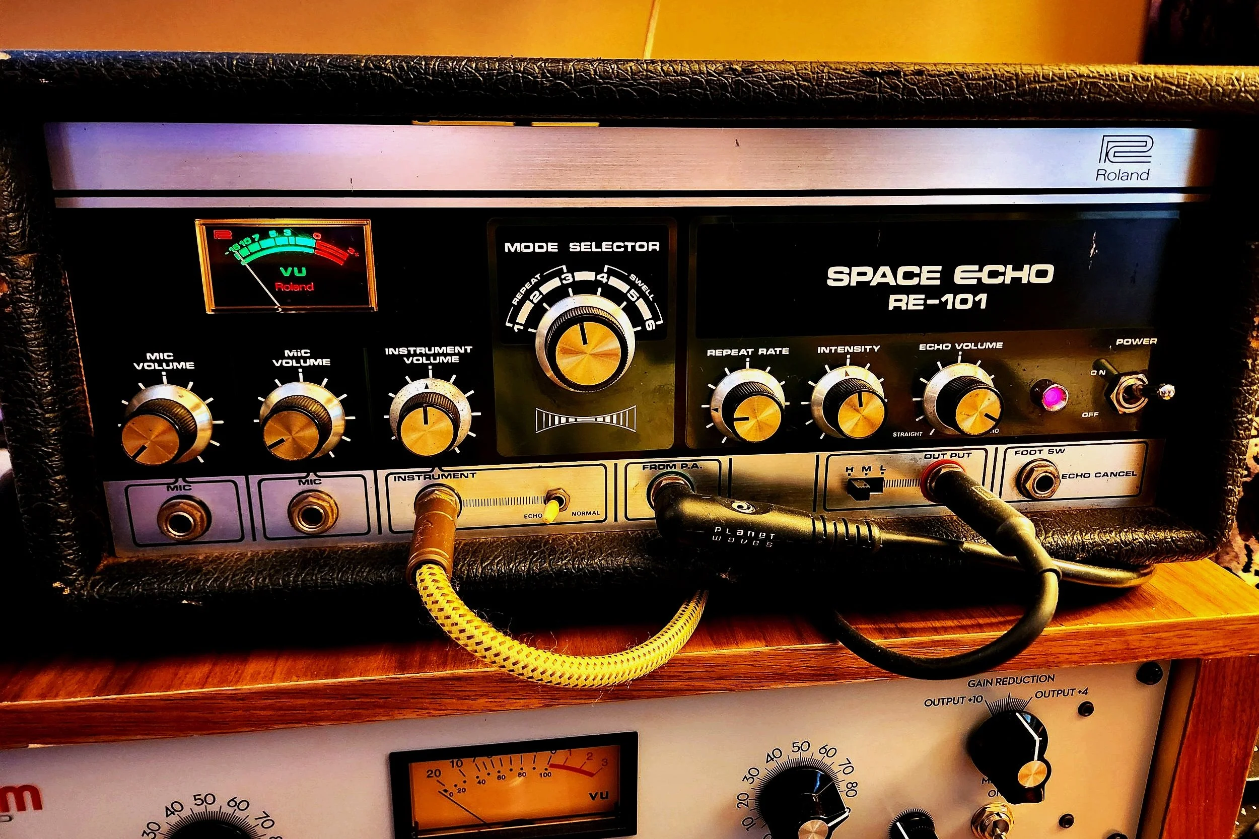 A vintage Roland Space Echo RE-101 tape delay effects unit on a wooden table, connected with audio cables and surrounded by other audio equipment.
