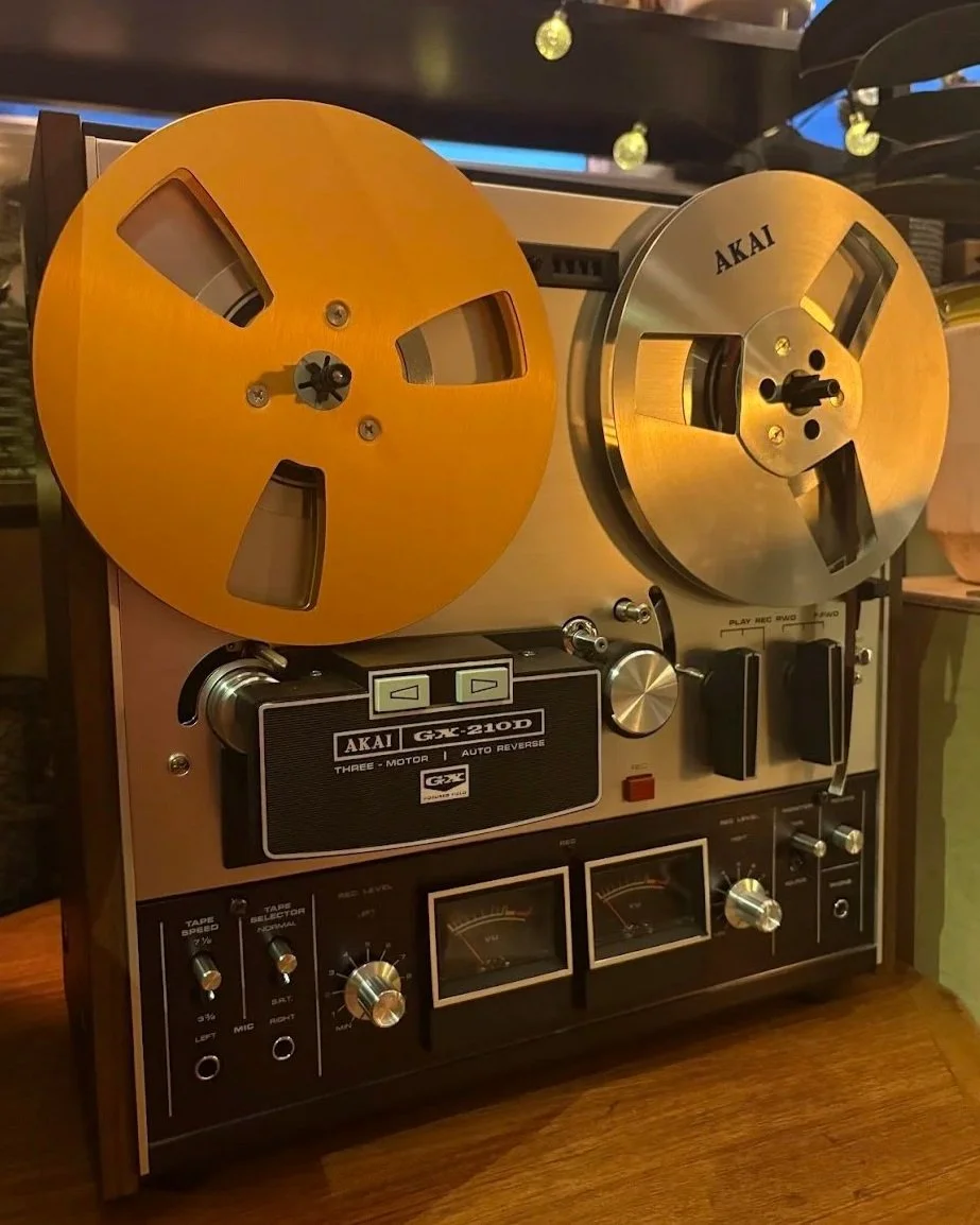 A vintage Akai reel-to-reel tape recorder with two large metal reels, one empty, on a wooden surface.