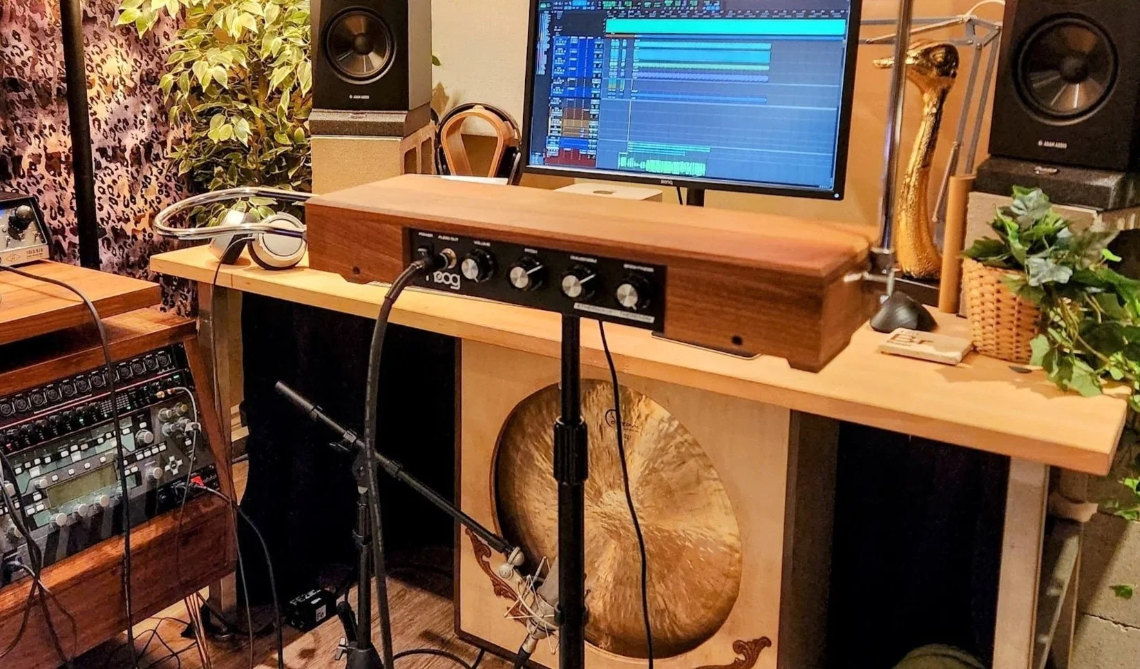 A music recording studio with a computer monitor displaying editing software, various audio equipment, headphones on a wooden stand, potted plants, a microphone, and a saxophone in the background.