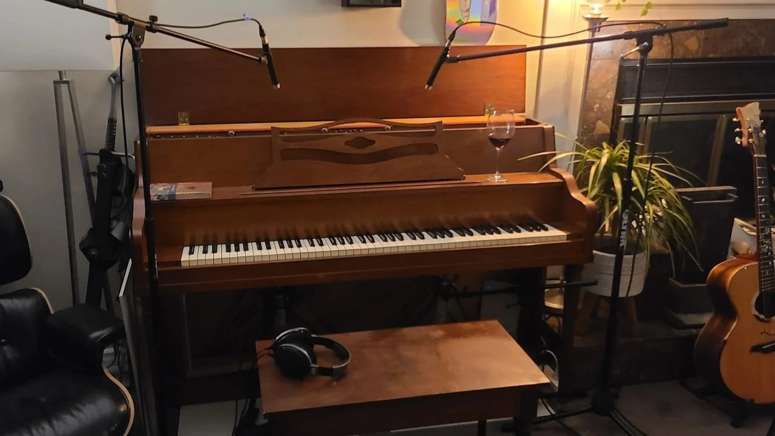 A brown upright piano with a glass of red wine on top, surrounded by musical equipment including microphones, an acoustic guitar, and headphones in a cozy room.