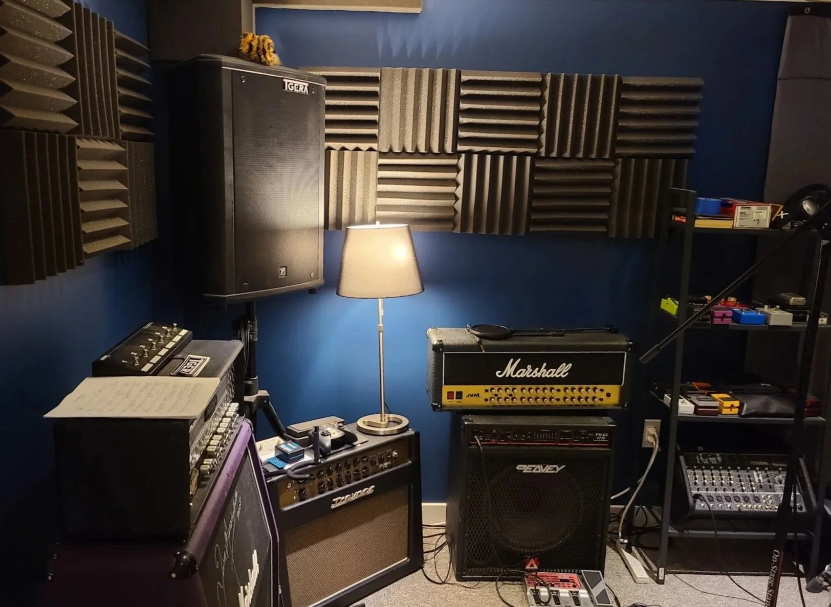 Music studio with acoustic foam panels, a Marshall guitar amplifier, various effects pedals, a lamp, and audio equipment.