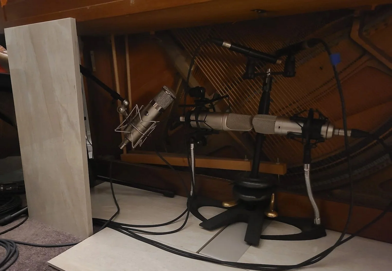 Inside a recording studio with two large condenser microphones mounted on stands in front of a grand piano, with cables running across the floor.