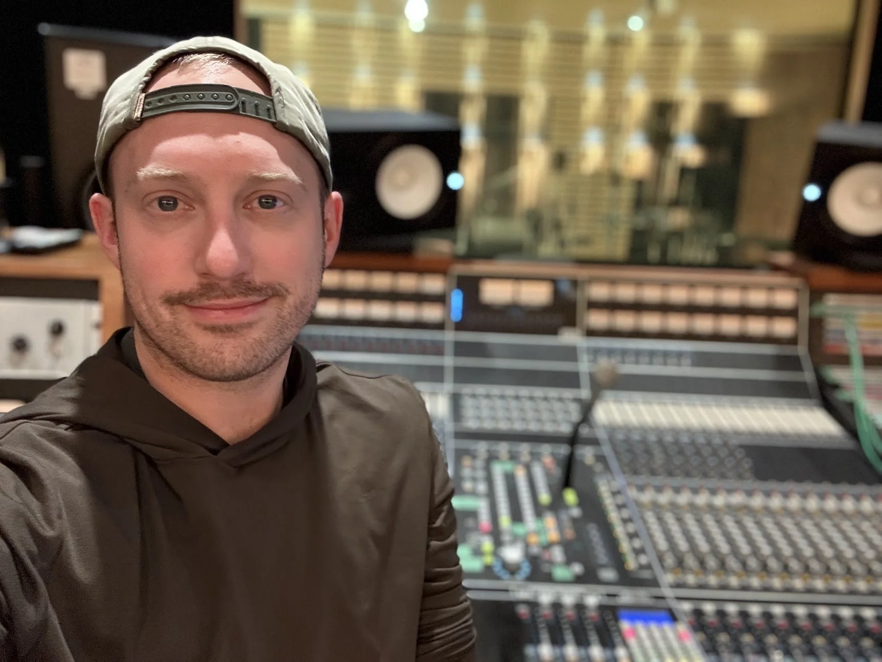 A man taking a selfie in a recording studio, with a large mixing console and studio monitors in the background.
