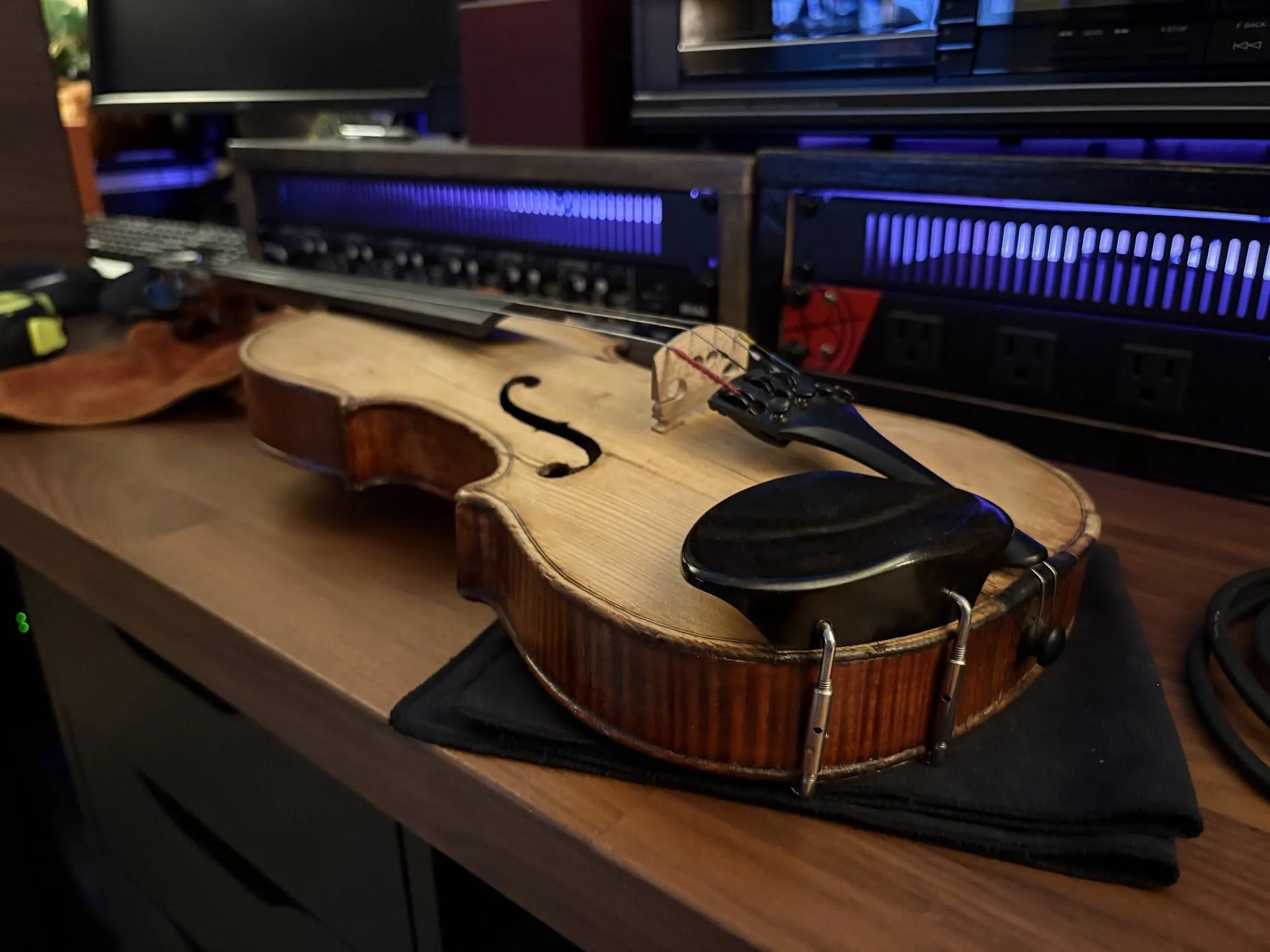 A violin lying on a desk with a black chin rest attached, in front of audio equipment with blue lighting.