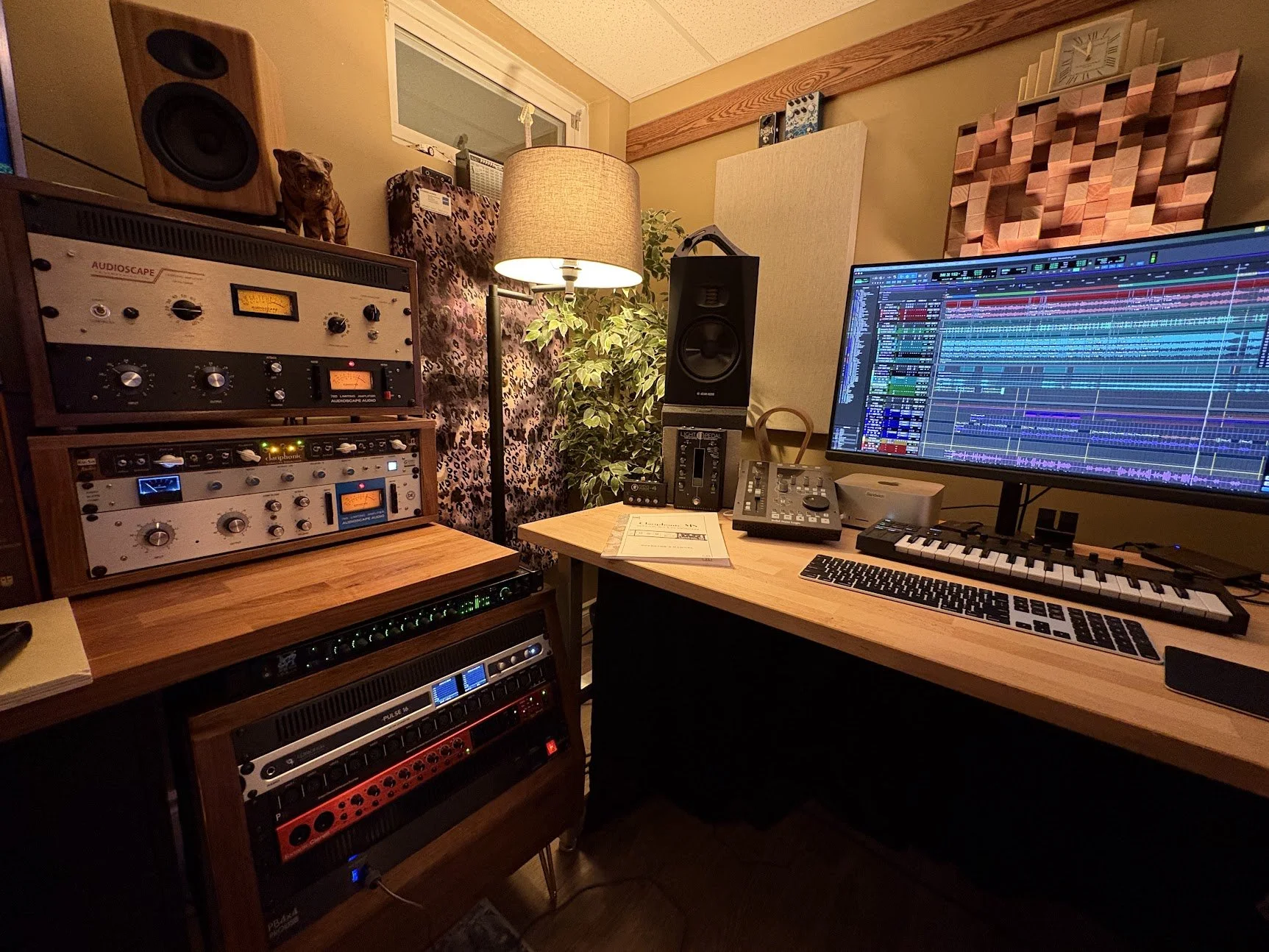 A music production studio with a computer monitor displaying digital audio workstation software, various audio equipment, speakers, a keyboard, a lamp, and decorative items including a leopard print soundproofing panel, a plant, a clock, and a tiger figurine.
