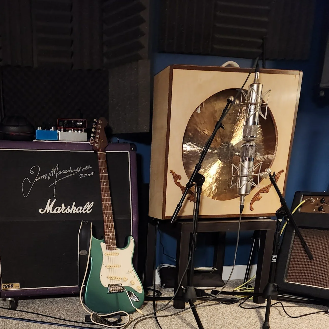 Music recording studio with a green electric guitar, Marshall amplifier, large golden gong, professional microphone on a stand, and various audio equipment.