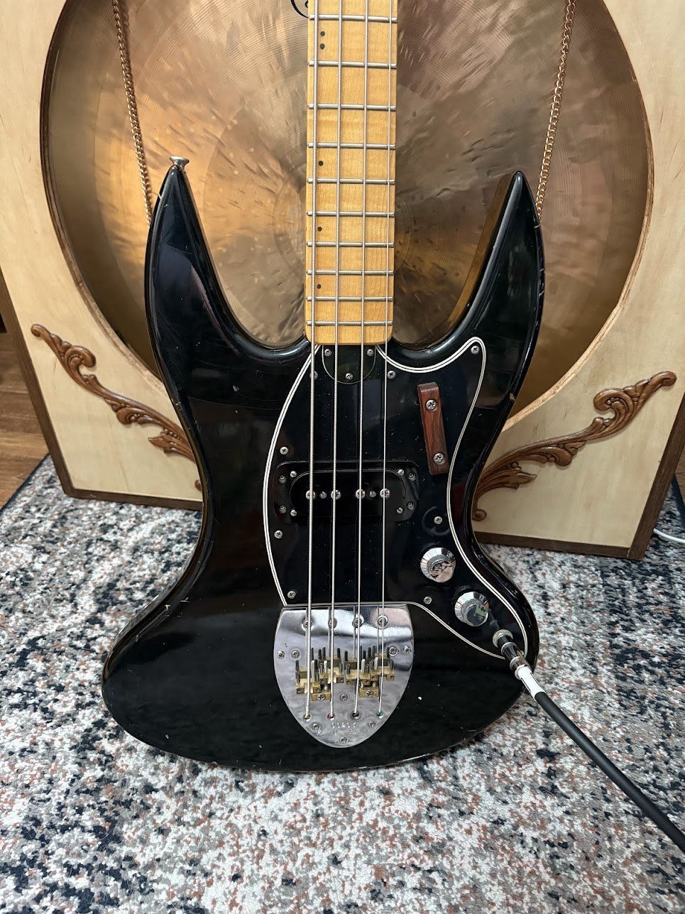 A black electric bass guitar with a distinctive shape, placed on a carpet in front of a large guitar-shaped speaker with a shiny, textured interior surface.