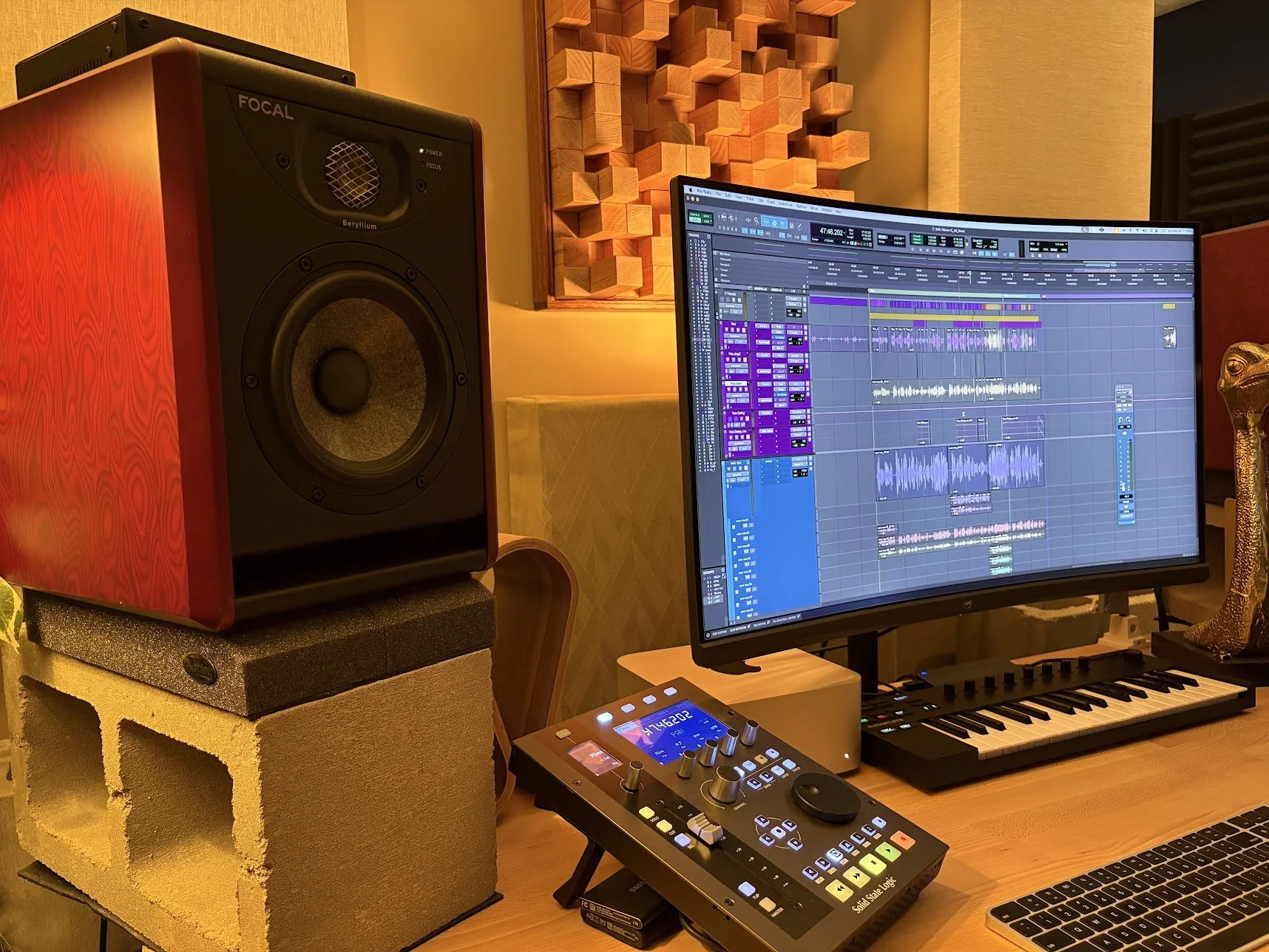 A music studio workstation with a computer monitor displaying music production software, a MIDI keyboard, a control surface, studio monitor speakers, acoustic panels on the wall, and a keyboard on a wooden desk.