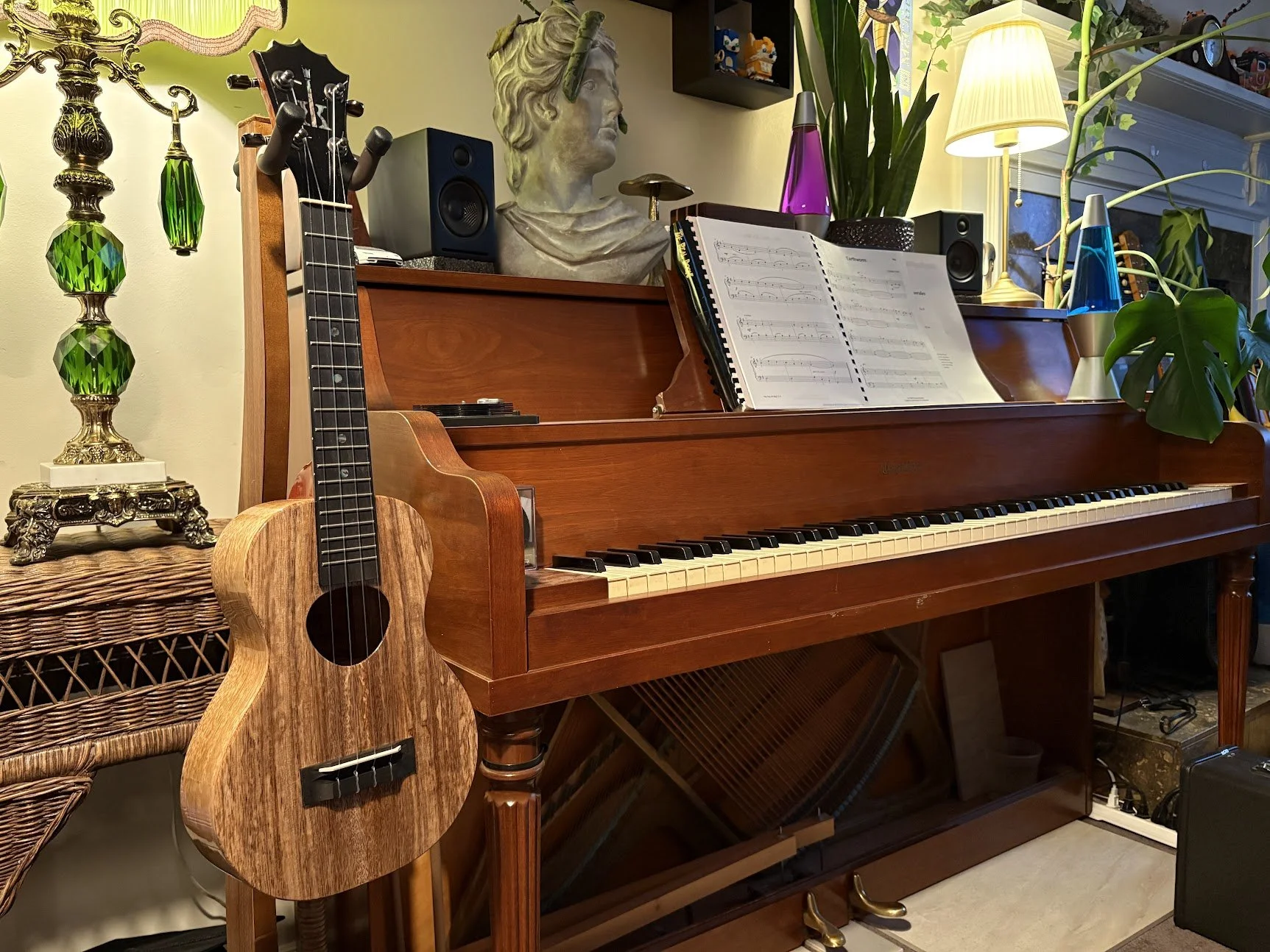 A wooden piano with sheet music on top, an acoustic guitar leaning against it, and a decorative lamp with green and purple glass accessories surrounded by plants and figurines in a cozy indoor setting.
