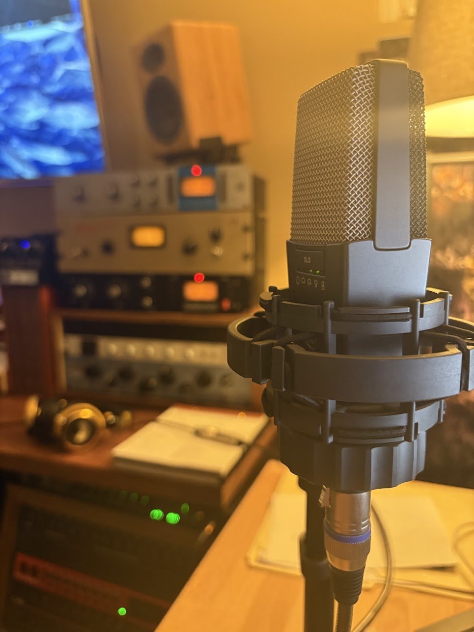 A studio microphone on a stand in a recording booth with audio equipment, a computer screen, and a music notebook in the background.