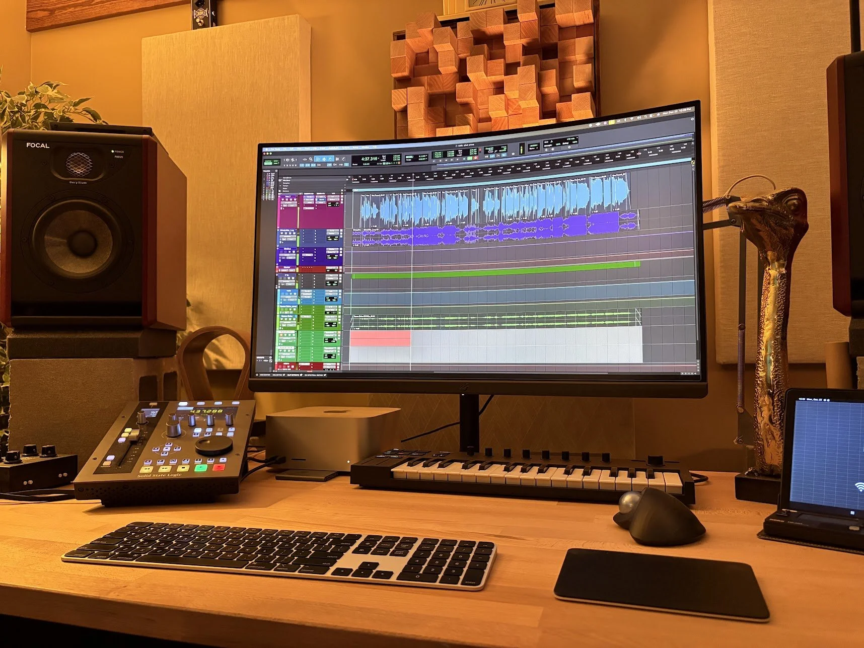 A music production workspace featuring a computer monitor displaying digital audio workstation software, studio monitor speakers, a MIDI keyboard, a DJ controller, a tablet, and various audio equipment on a wooden desk in a soundproof studio with aco