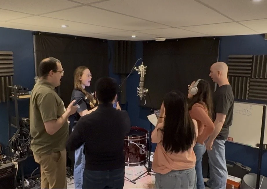 Six people in a recording studio with music equipment and a microphone, engaged in a discussion or recording session.