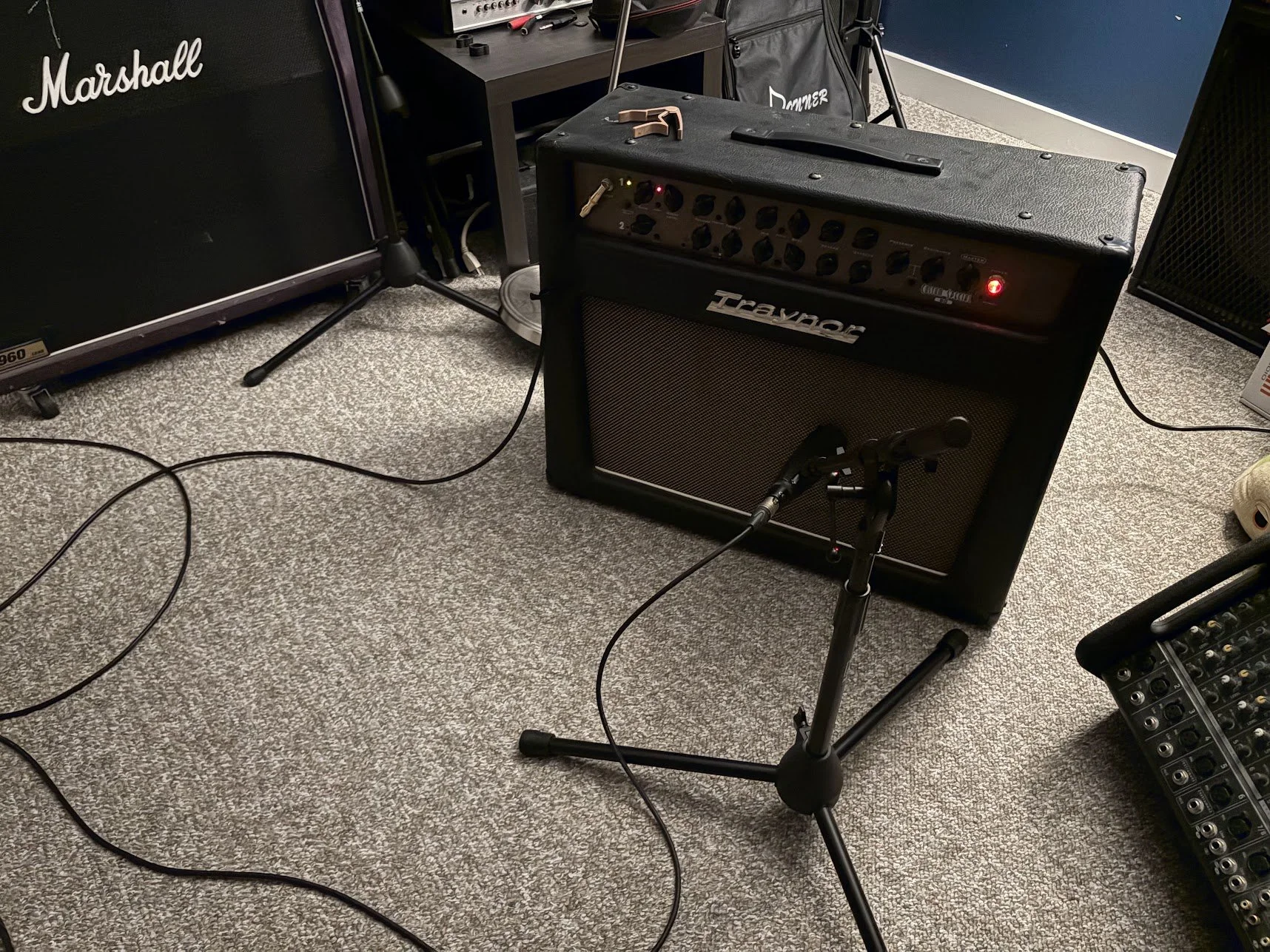 A music practice space with a guitar amplifier, microphone on a stand, and various audio equipment including a Marshall speaker, a ZT guitar amplifier, and a small mixing console on a carpeted floor.