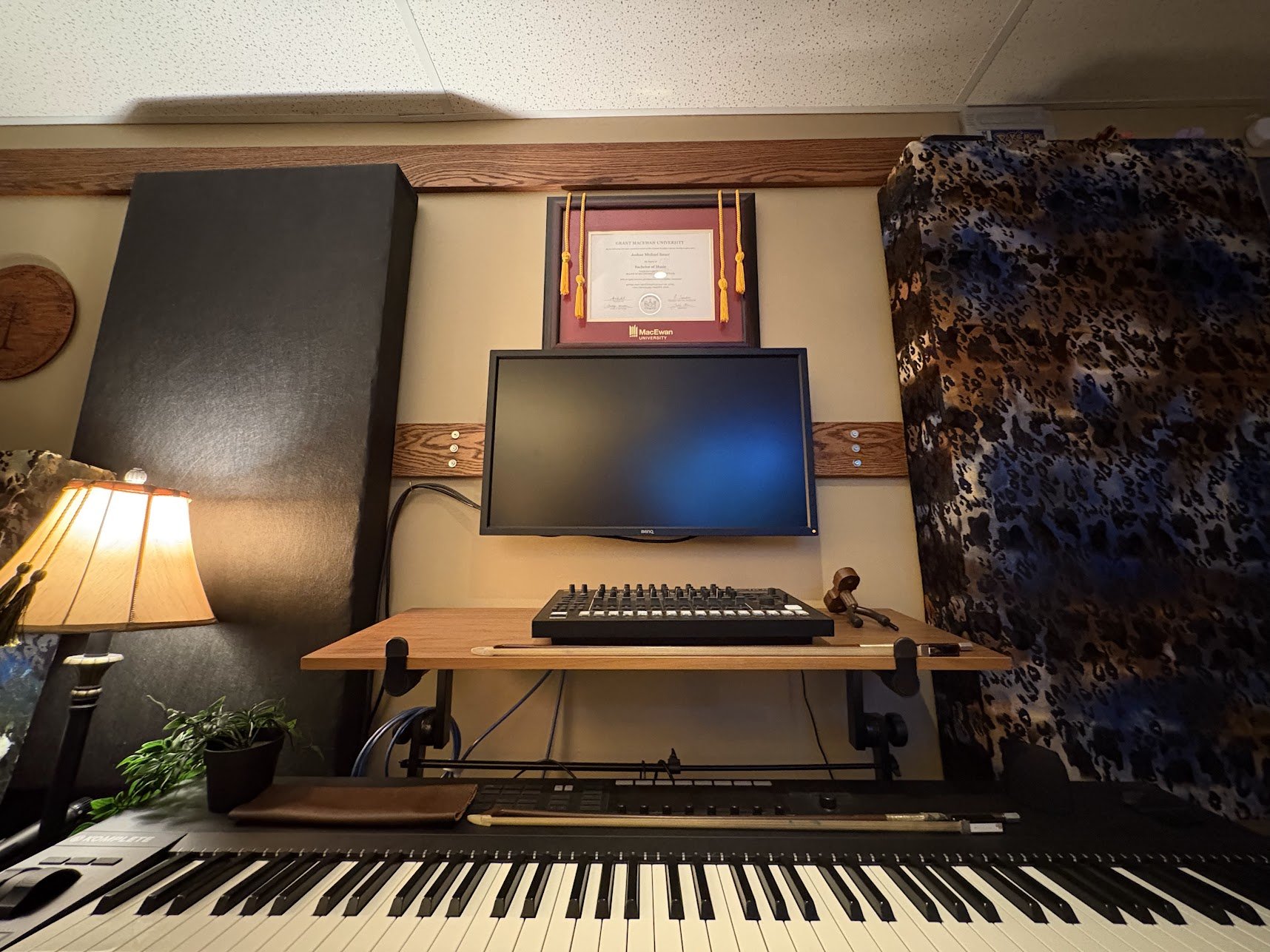 A music studio setup with a keyboard in the foreground, a small mixing console on a wooden desk, a monitor, and a framed diploma on the wall above the monitor. There is a lamp on the left side, a plant below, and a floral patterned curtain on the rig