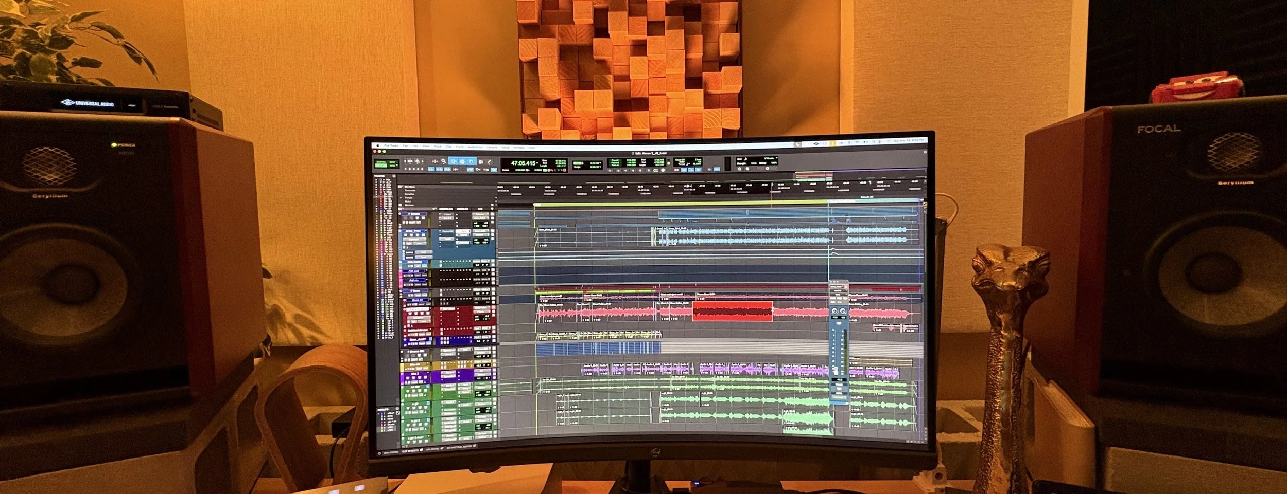 A music production studio with a large computer monitor displaying digital audio workstation software, surrounded by studio monitors on either side, a decorative sculpture resembling a lion's head on the right, and acoustic foam panels on the wall in