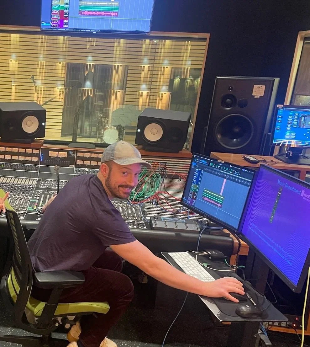 A man in a recording studio at a mixing console with two computer monitors, studio speakers, and a large window into the recording room.