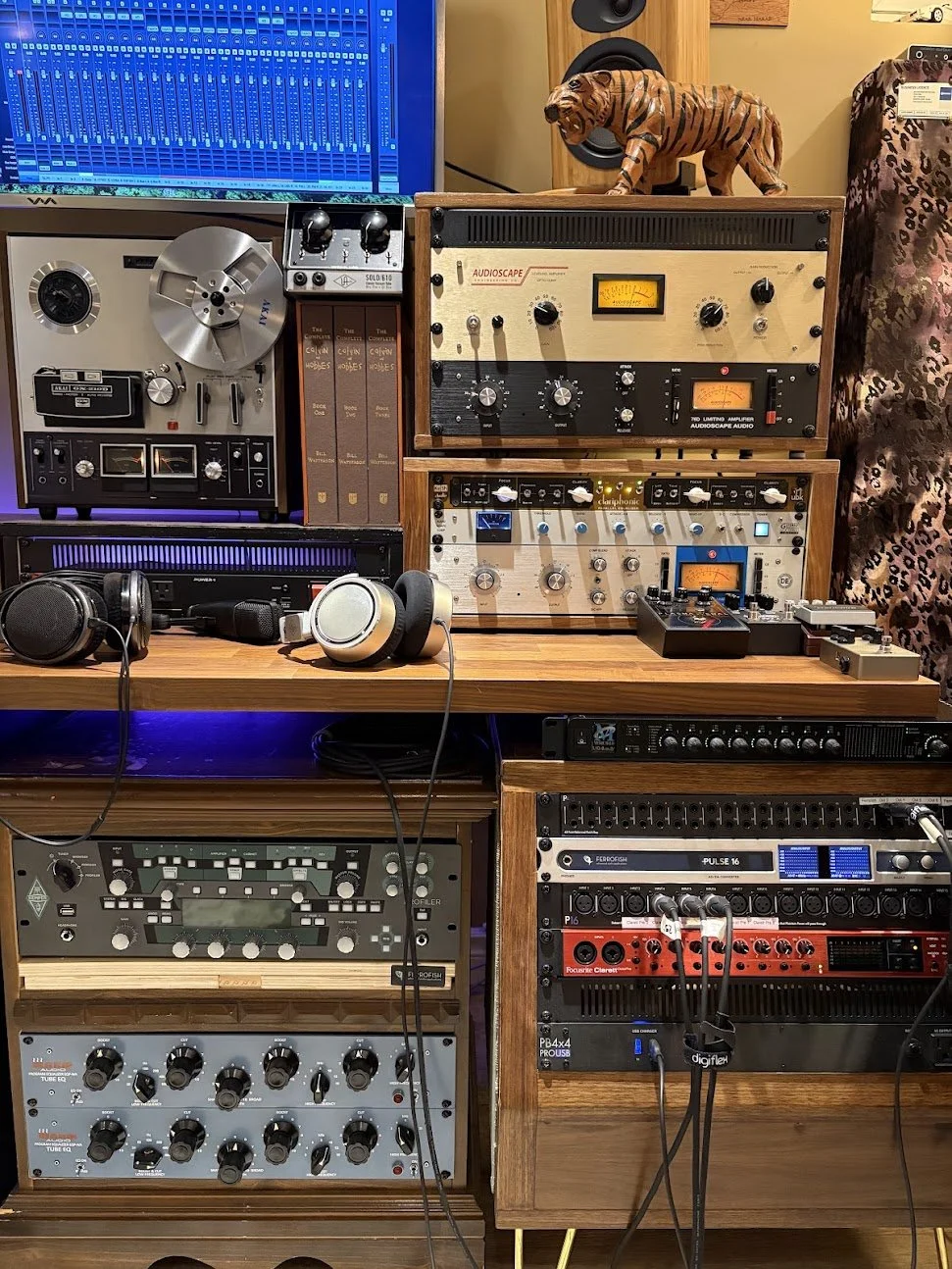 A music studio with vintage audio equipment, including a reel-to-reel tape recorder, analog amplifiers, and mixing gear. There are headphones on a wooden desk, and a small tiger figurine on top of an amplifier. A computer monitor displays a digital audio workstation, and other audio hardware is arranged systematically.
