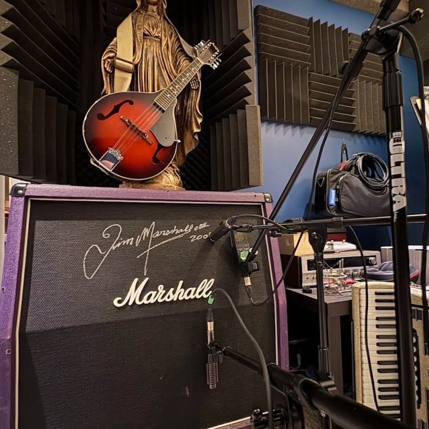 A music studio with a Marshall amplifier signed by Jim Marshall, an acoustic guitar hanging on a wooden statue, a microphone stand, an audio recorder, and soundproofing foam panels.