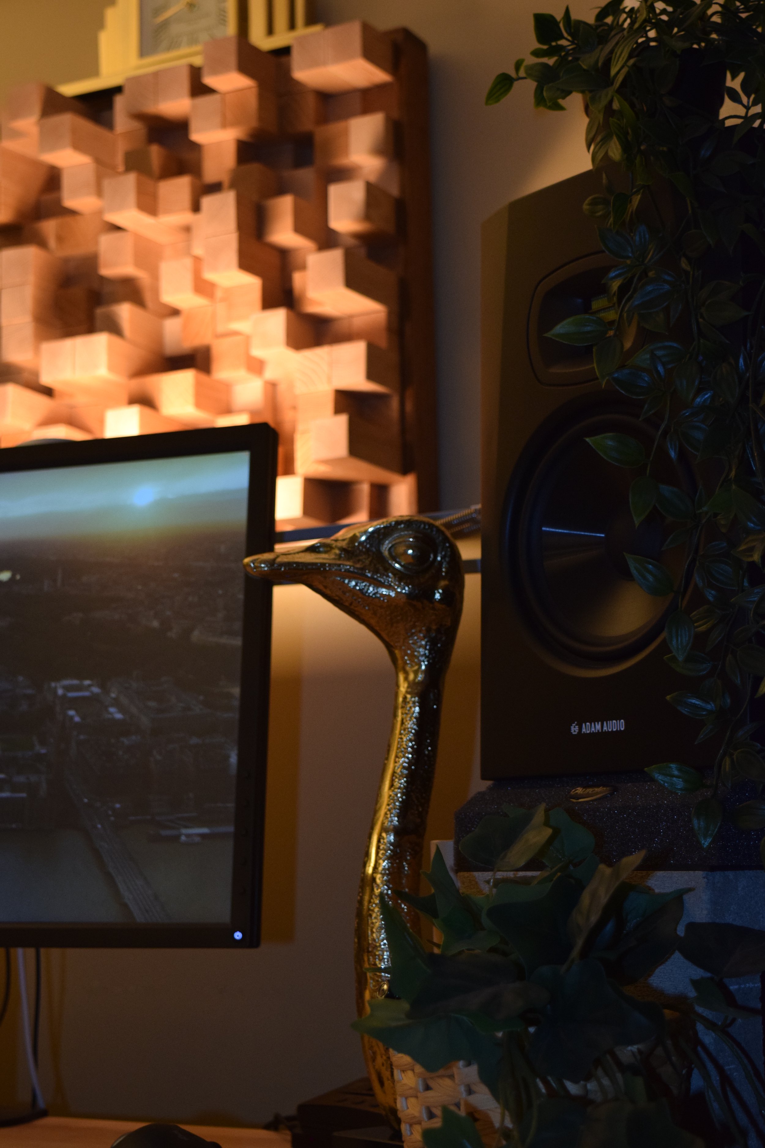 A computer monitor displaying a scene, a decorative wooden wall art piece, a green leafy plant, a gold-colored giraffe figurine, and an Adam Audio speaker.