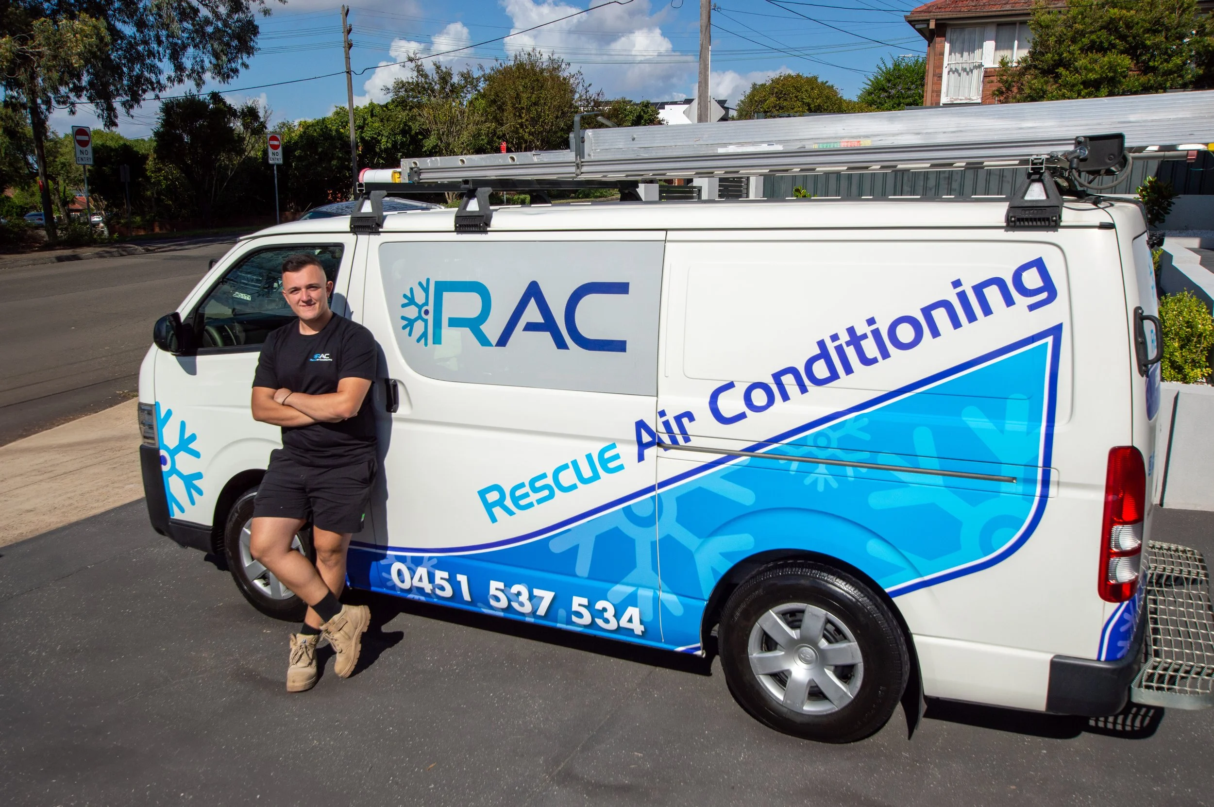 A young man leaning against a white rescue air conditioning service van parked on a street. The van displays the company logo, a snowflake symbol, and contact information. The man is smiling, wearing black shorts, a black t-shirt with a logo, boots, and has arms crossed. There are trees, utility poles, and houses in the background under a partly cloudy sky.