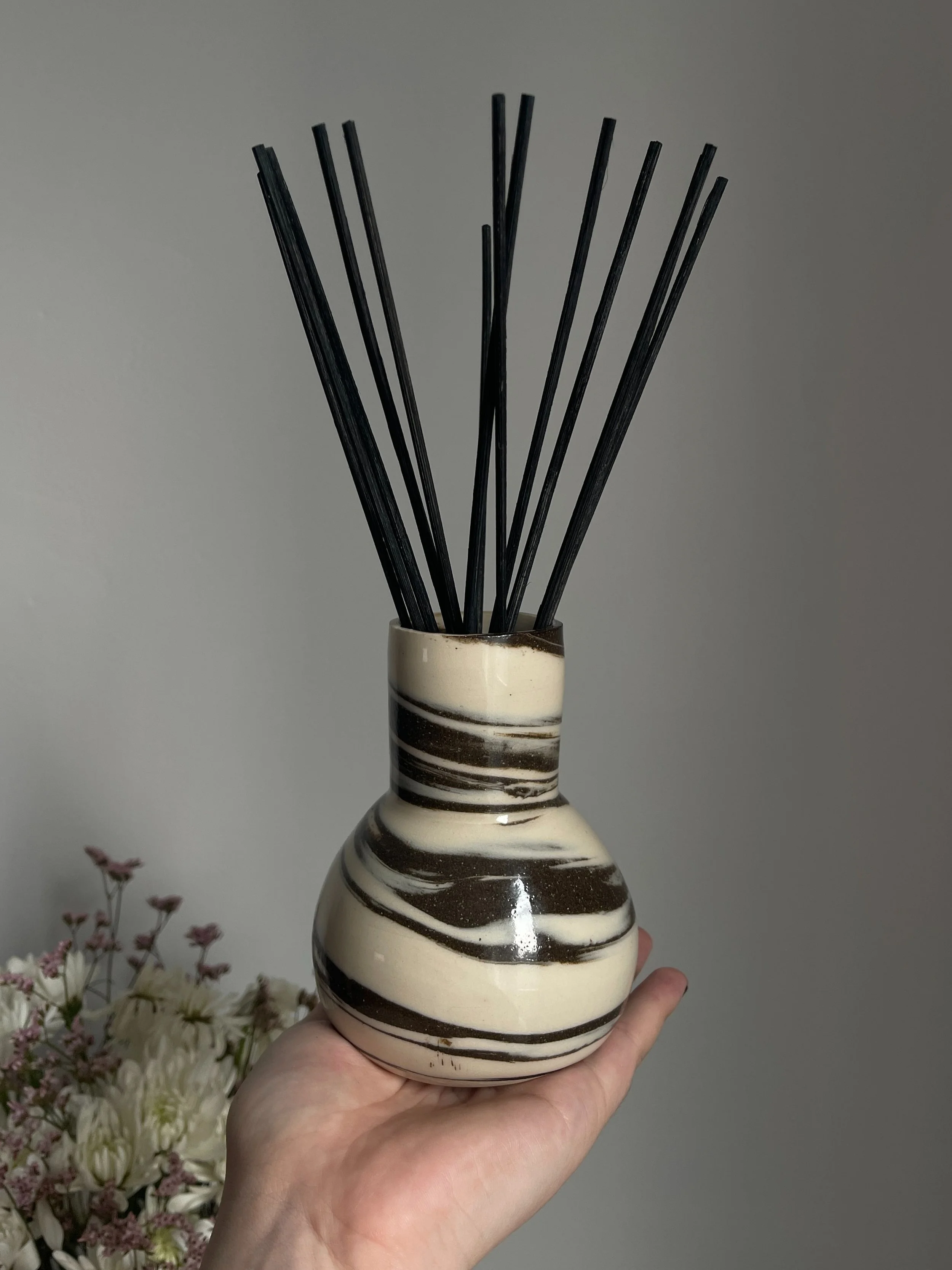 Hand holding a ceramic diffuser with black reeds in a swirled brown and white design, set against a gray background.