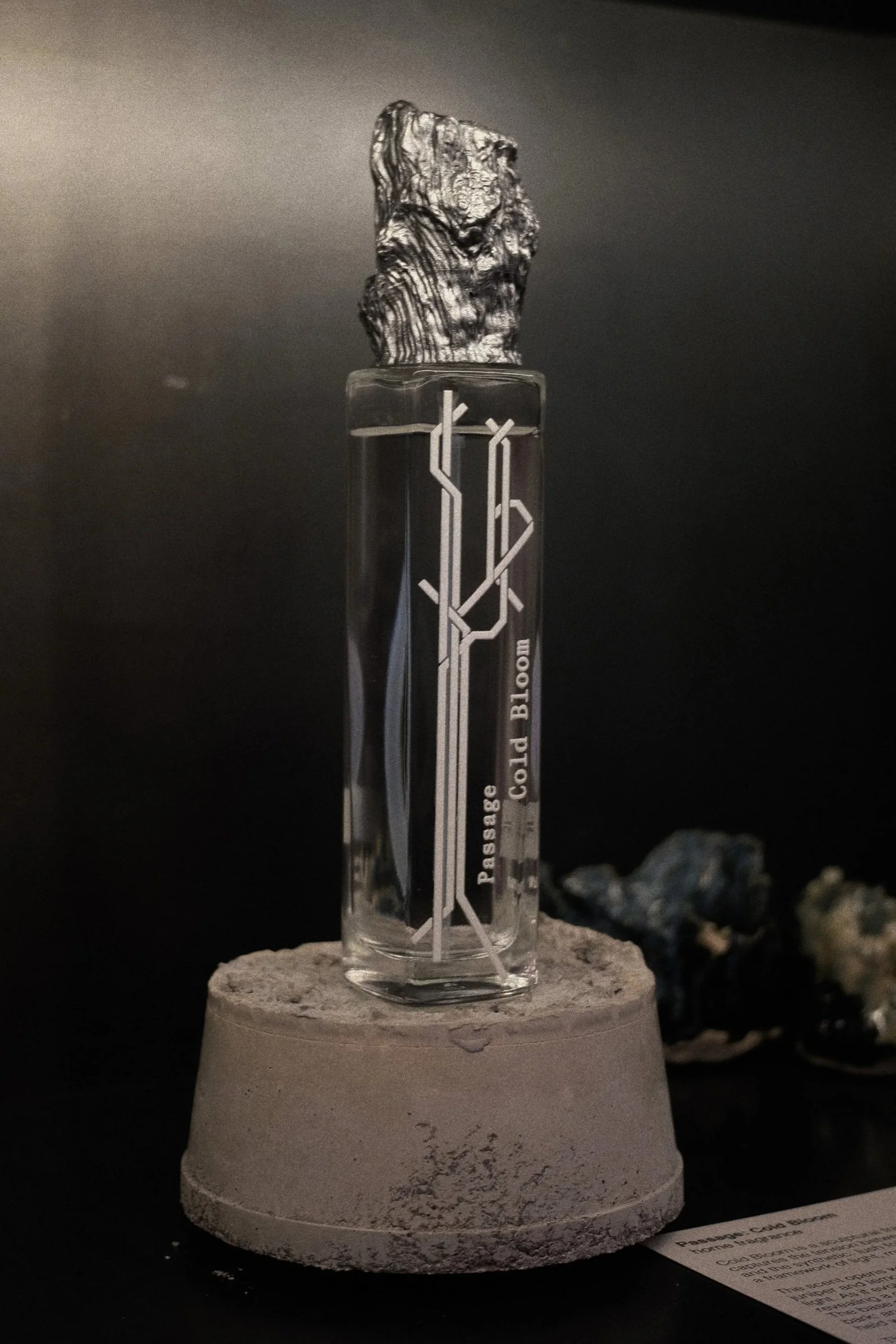 A glass container with the words 'Passed Cold Bloom' on it, placed on a concrete pedestal, with a piece of textured rock on top. The background is dark and plain.