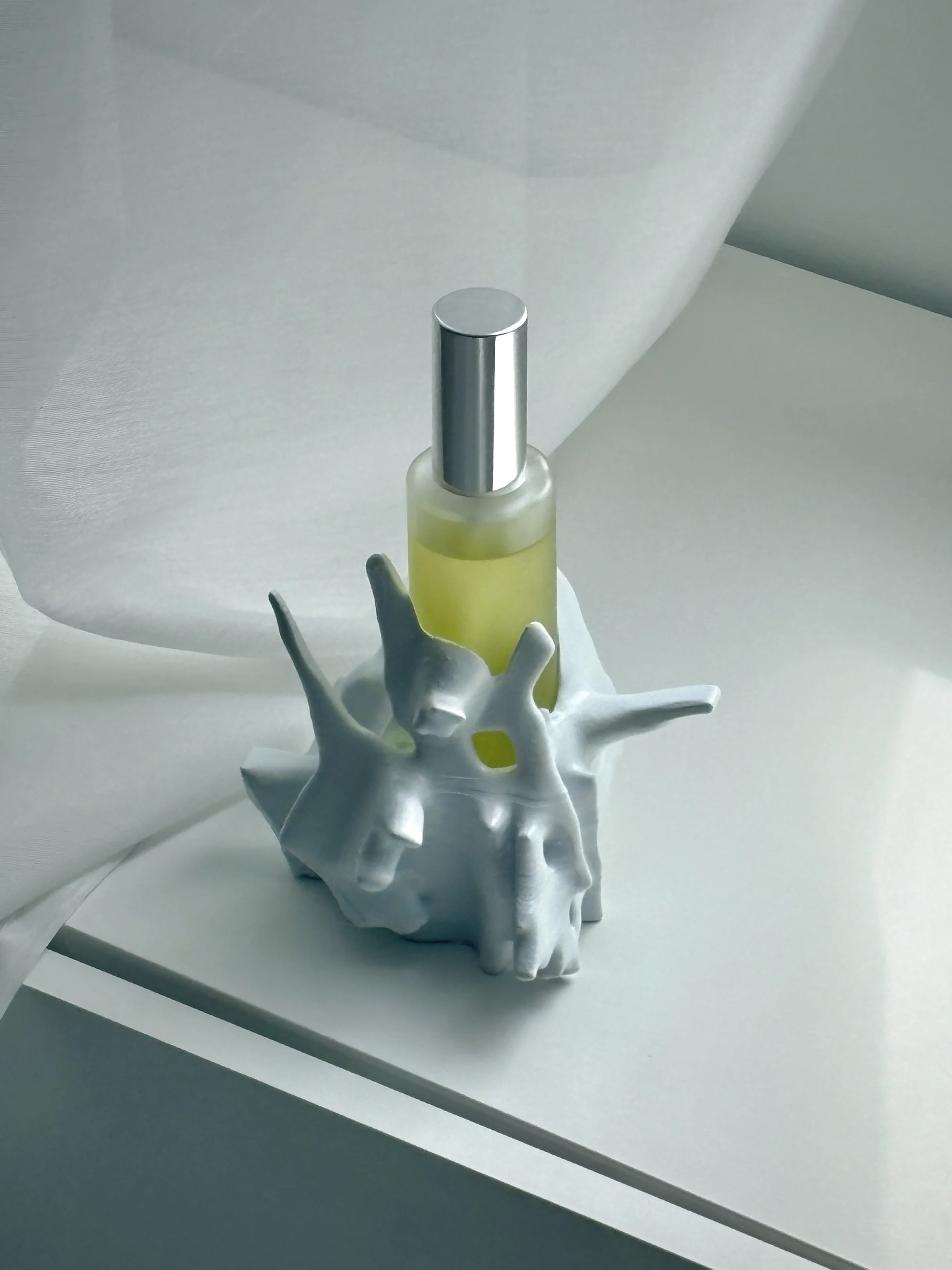 A bottle of yellow liquid with a silver cap, placed inside a white, abstract ceramic holder on a light surface, partially covered by translucent white fabric.