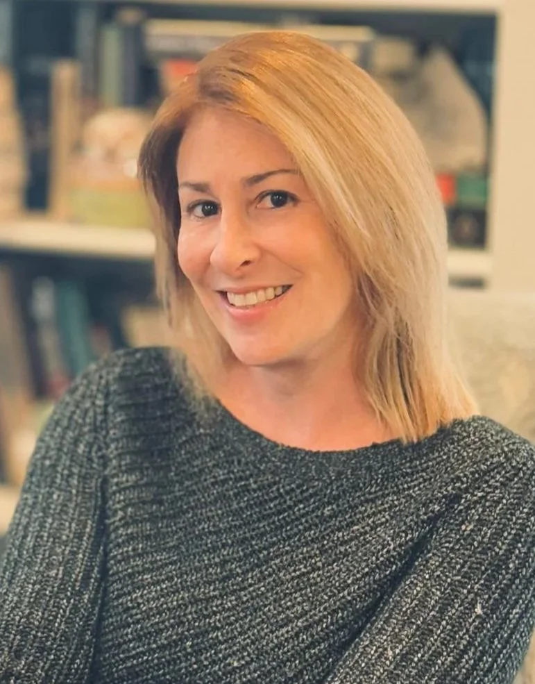 A woman with shoulder-length blond hair, wearing a gray textured sweater, smiling and looking at the camera, with a bookshelf in the background.