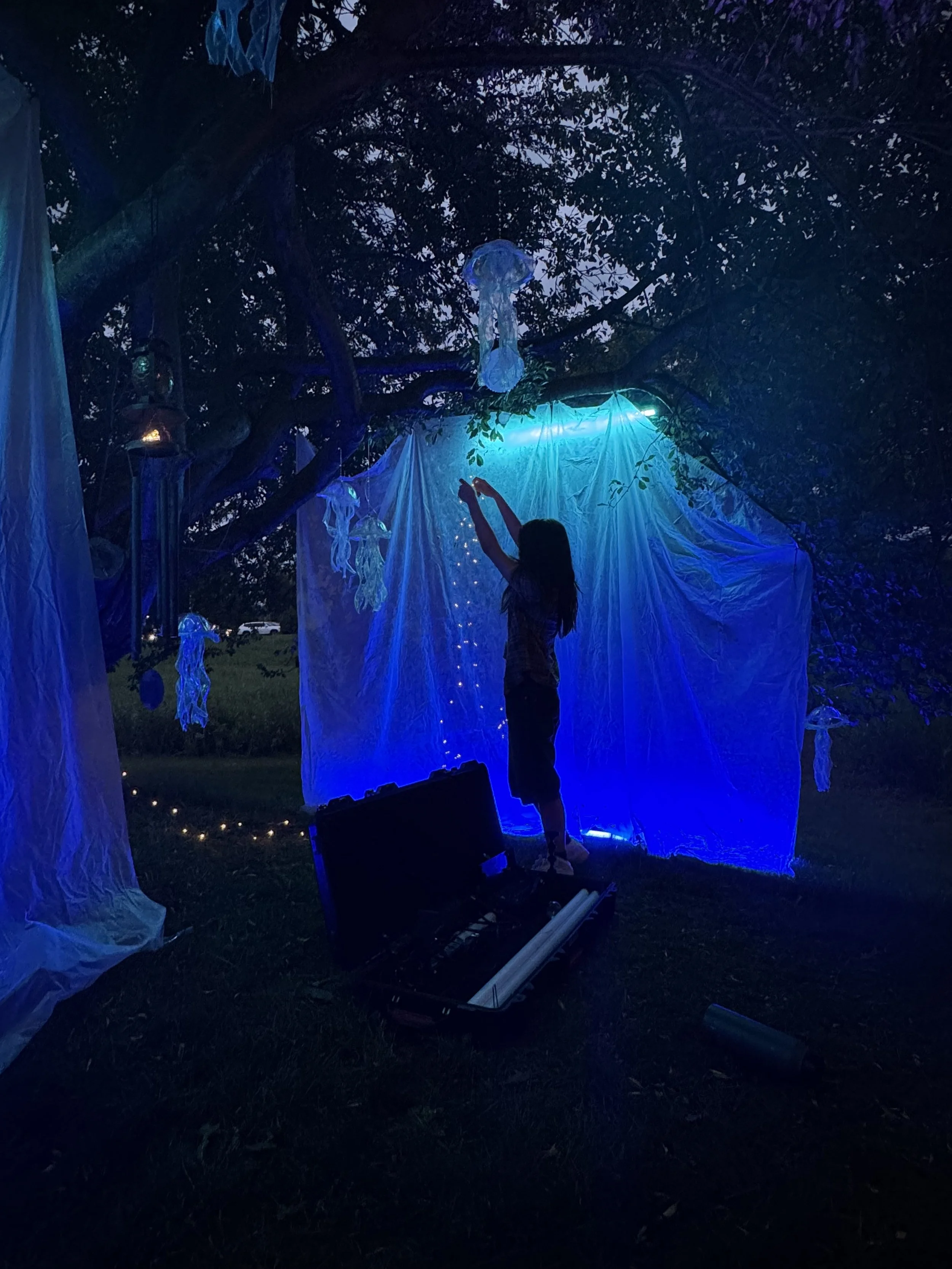 A person standing outdoors at dusk surrounded by hanging fabric decorations and illuminated jellyfish-shaped hanging ornaments with blue lighting.