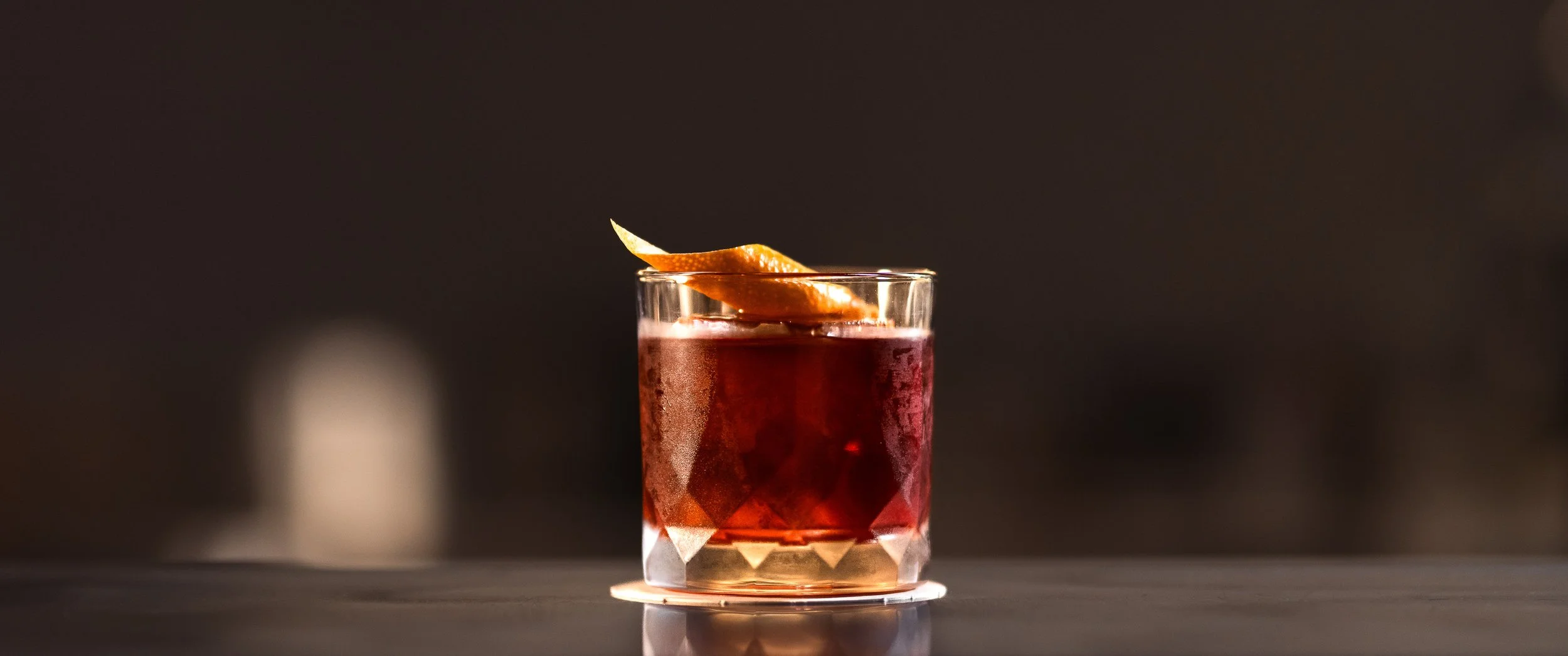 A glass of dark red cocktail with a lemon peel garnish on top, placed on a reflective surface with a blurred dark background. Brisbane by Talon Media.