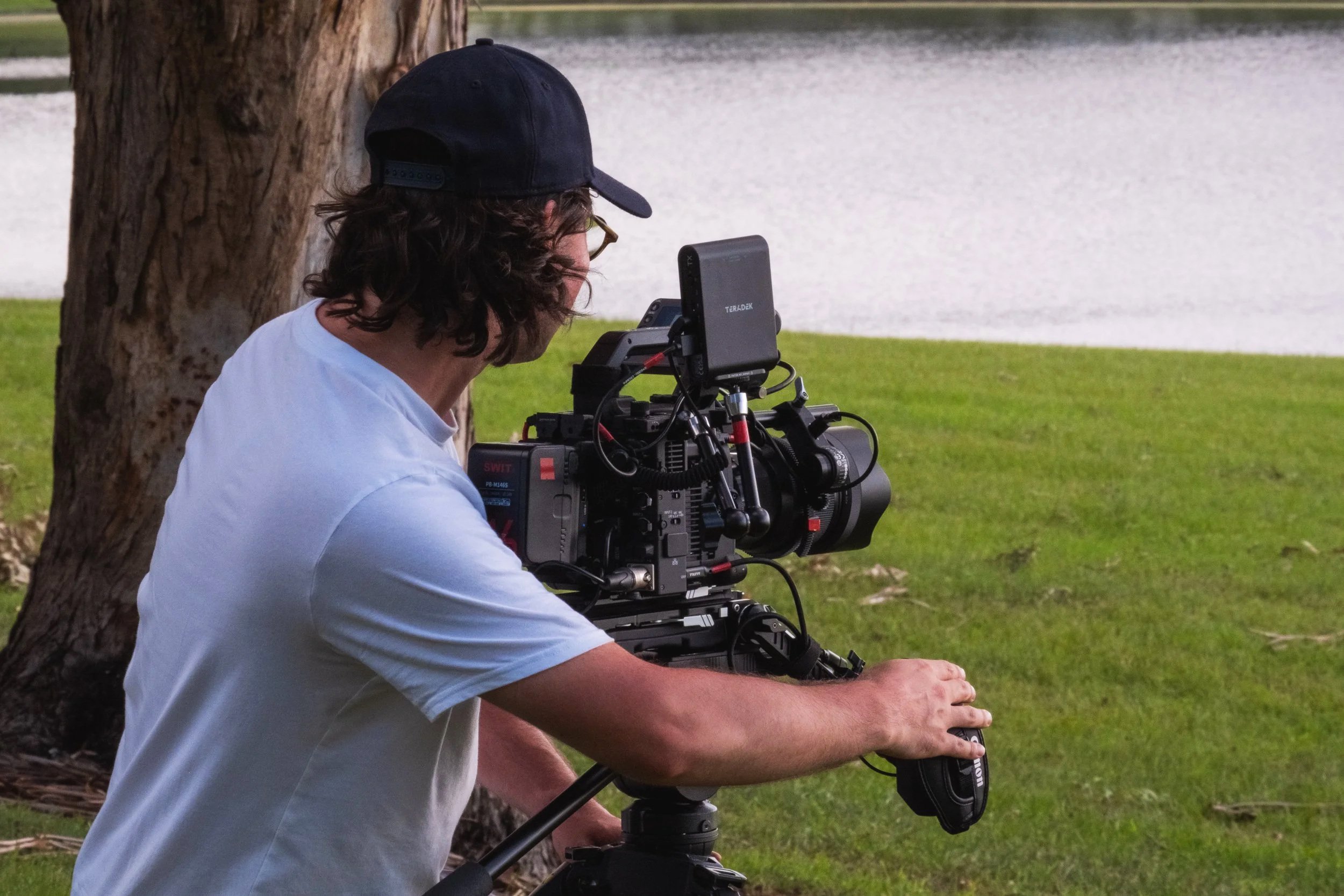 Hamish Powell from Talon Media wearing a white t-shirt and a black cap uses a professional film camera outdoors near a tree, with a lake and grassy area in the background.
