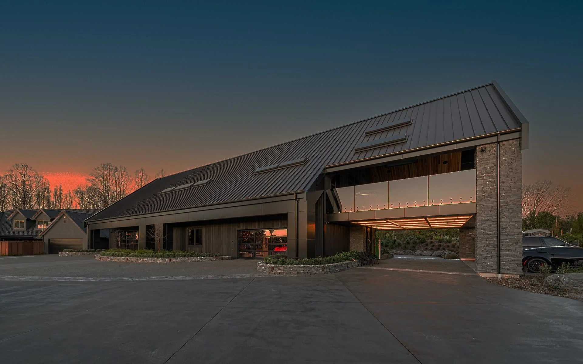 Modern building with a sloped metal roof at sunset, surrounded by trees and a paved driveway.