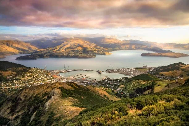 A panoramic view of a bay with a shoreline, boats, and a nearby town surrounded by green hills and mountains under a partly cloudy sky.
