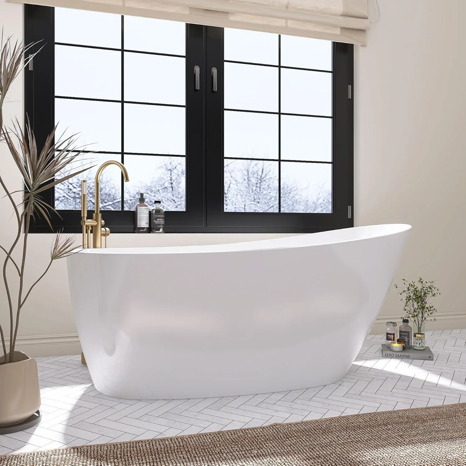 Modern bathroom with a white freestanding bathtub next to a large black-framed window with snow-covered trees outside. There are gold fixtures on the tub, potted plants, and various bottles of bath products.