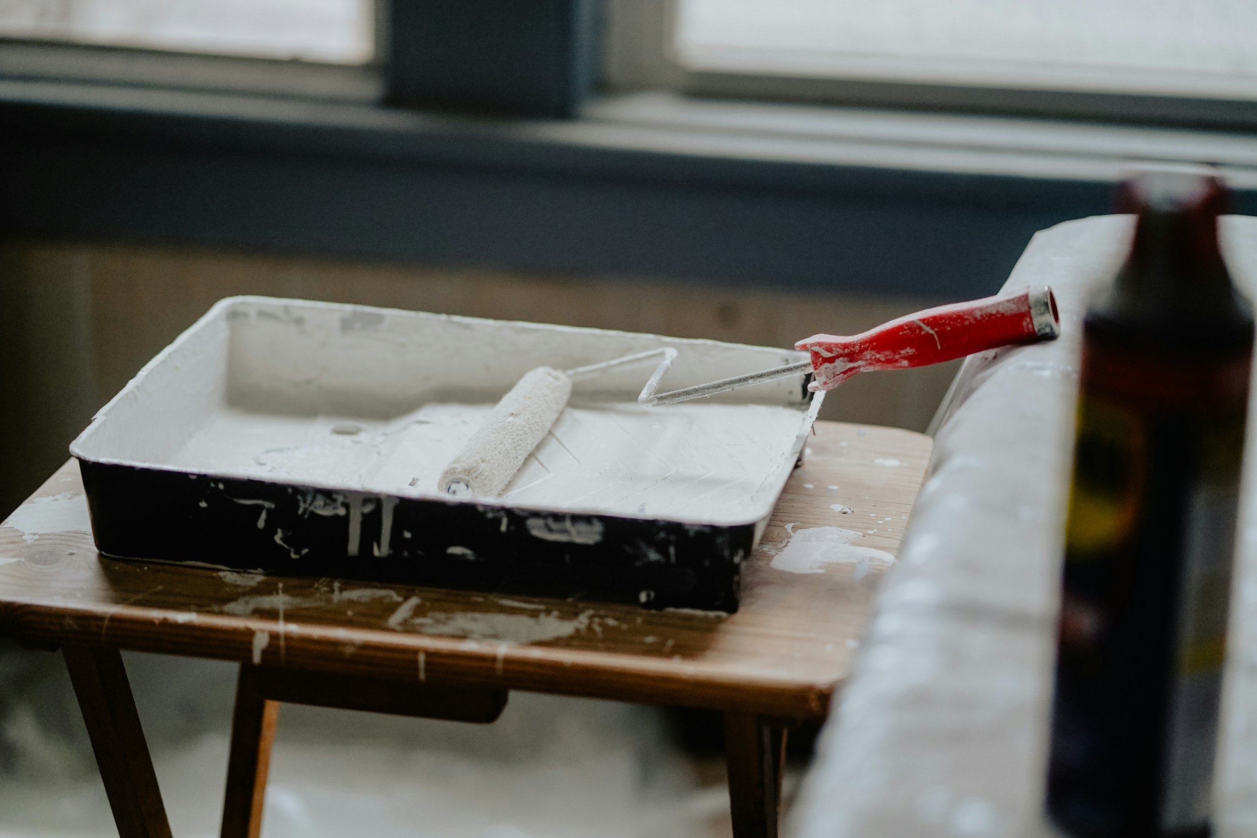 Paint tray with white paint, roller, paintbrush, and an open can of paint on a wooden table near a window.