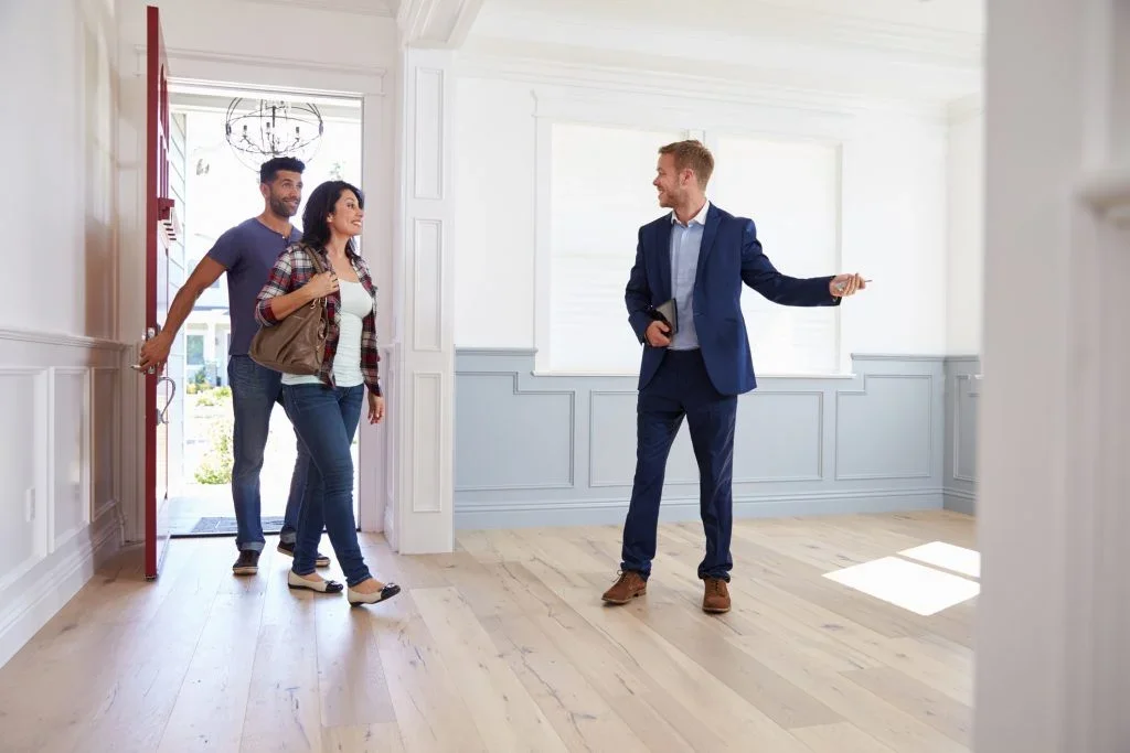 A real estate agent showing a house to a couple inside a bright, empty room with large windows and light-colored wooden floors.