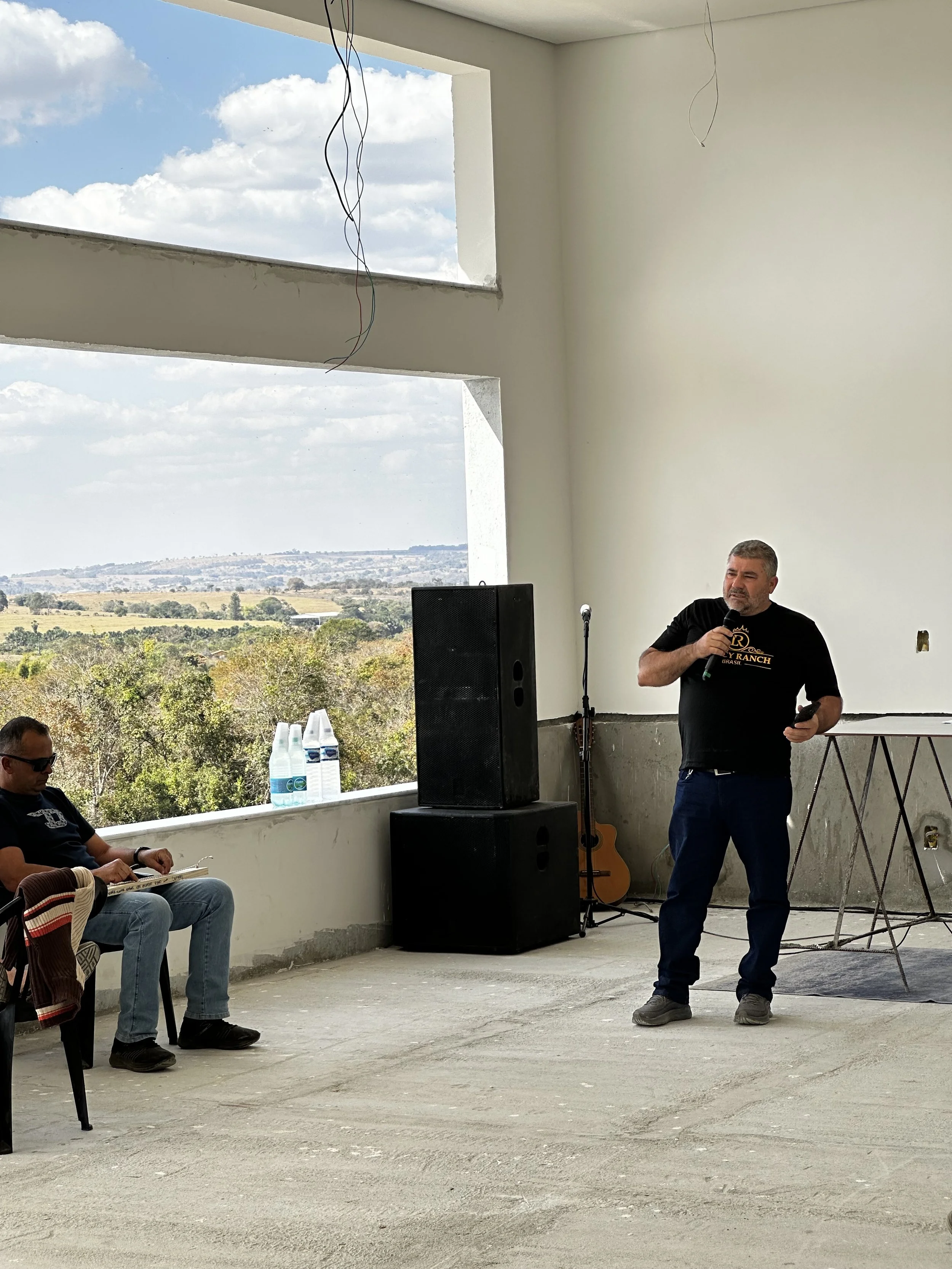 A man giving a speech in a room with large open windows showing a landscape of trees and hills, with some wires hanging from the ceiling, and a seated man listening.