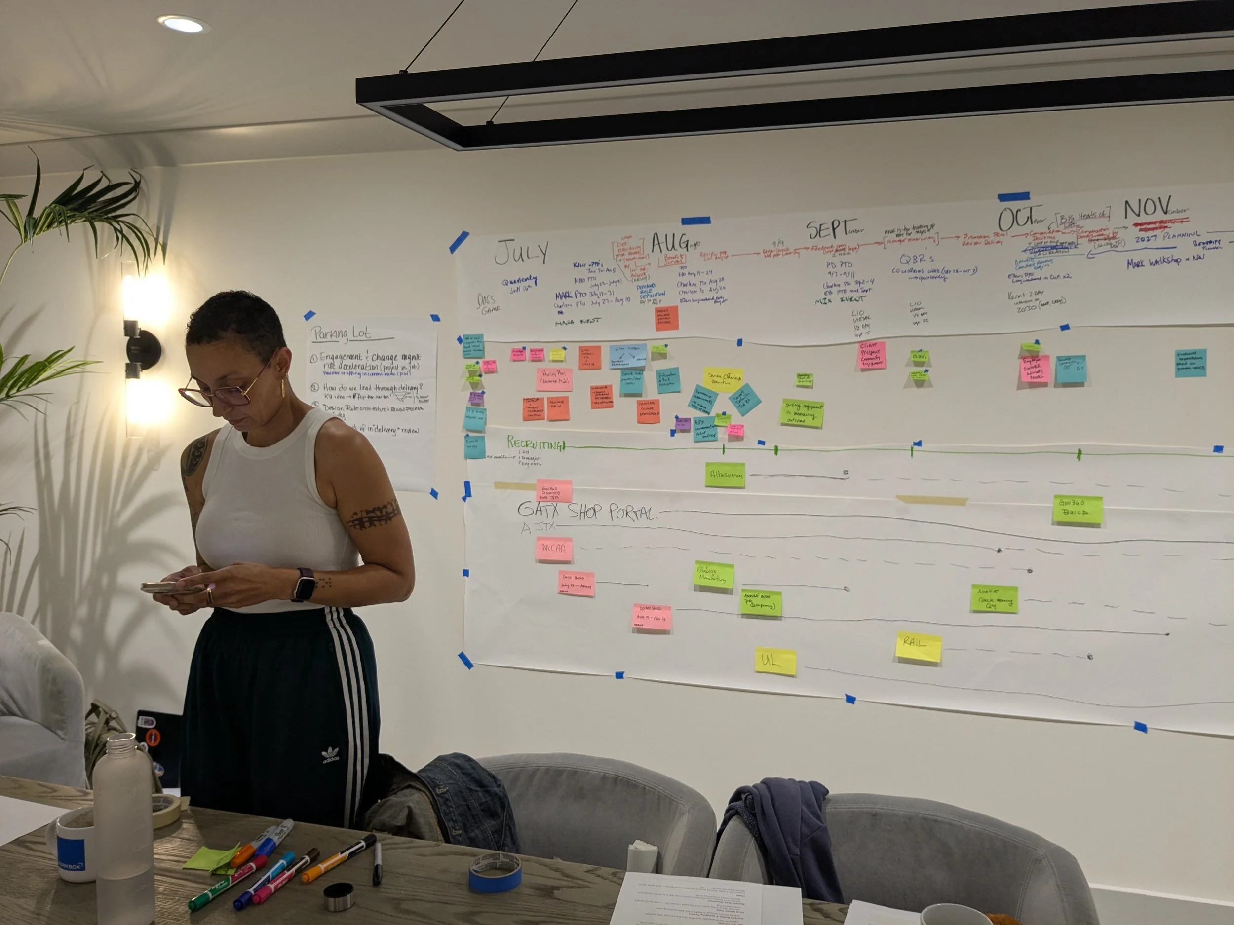 A woman wearing glasses and a tank top stands in front of a wall covered with colorful sticky notes and handwritten notes, with a table in the foreground holding markers, paper, and bottles.