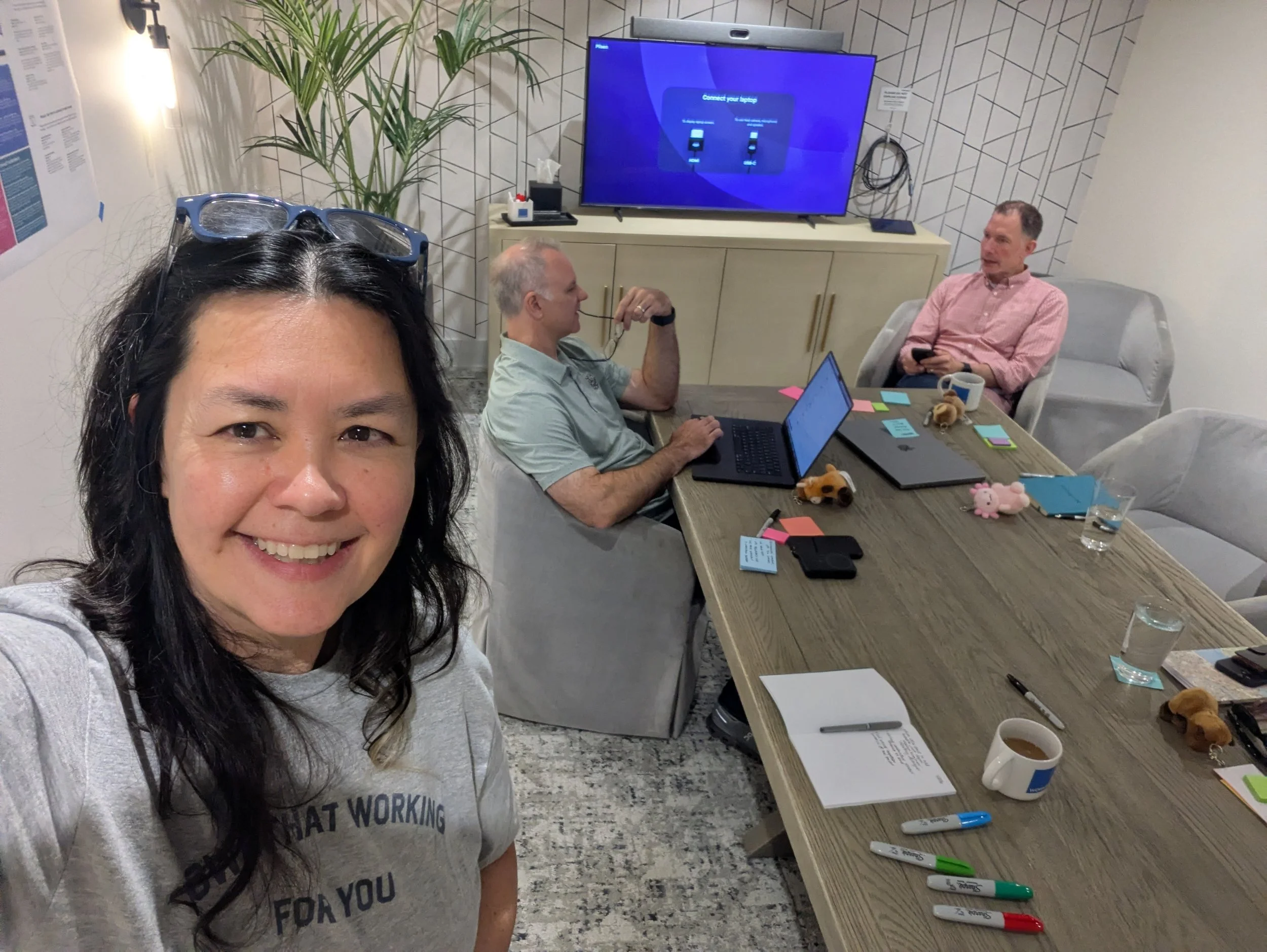 A woman with black hair and glasses on her head taking a selfie in a conference room. Behind her, two men are sitting at a table with a laptop, notebooks, and pens, engaged in a discussion. The table also has snacks, sticky notes, a coffee mug, and a container of tissues. A large TV screen on the wall in the background displays a disconnected screen, and there are decorative plants and a wall with a geometric pattern.
