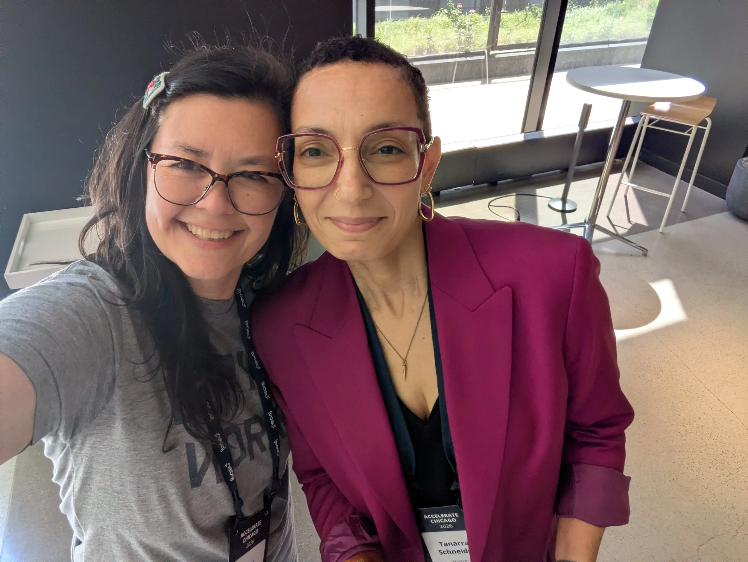 2 women smiling at the camera in selfie mode. Both have glasses, 1 is wearing a gray t-shirt and the other a magenta blazer.