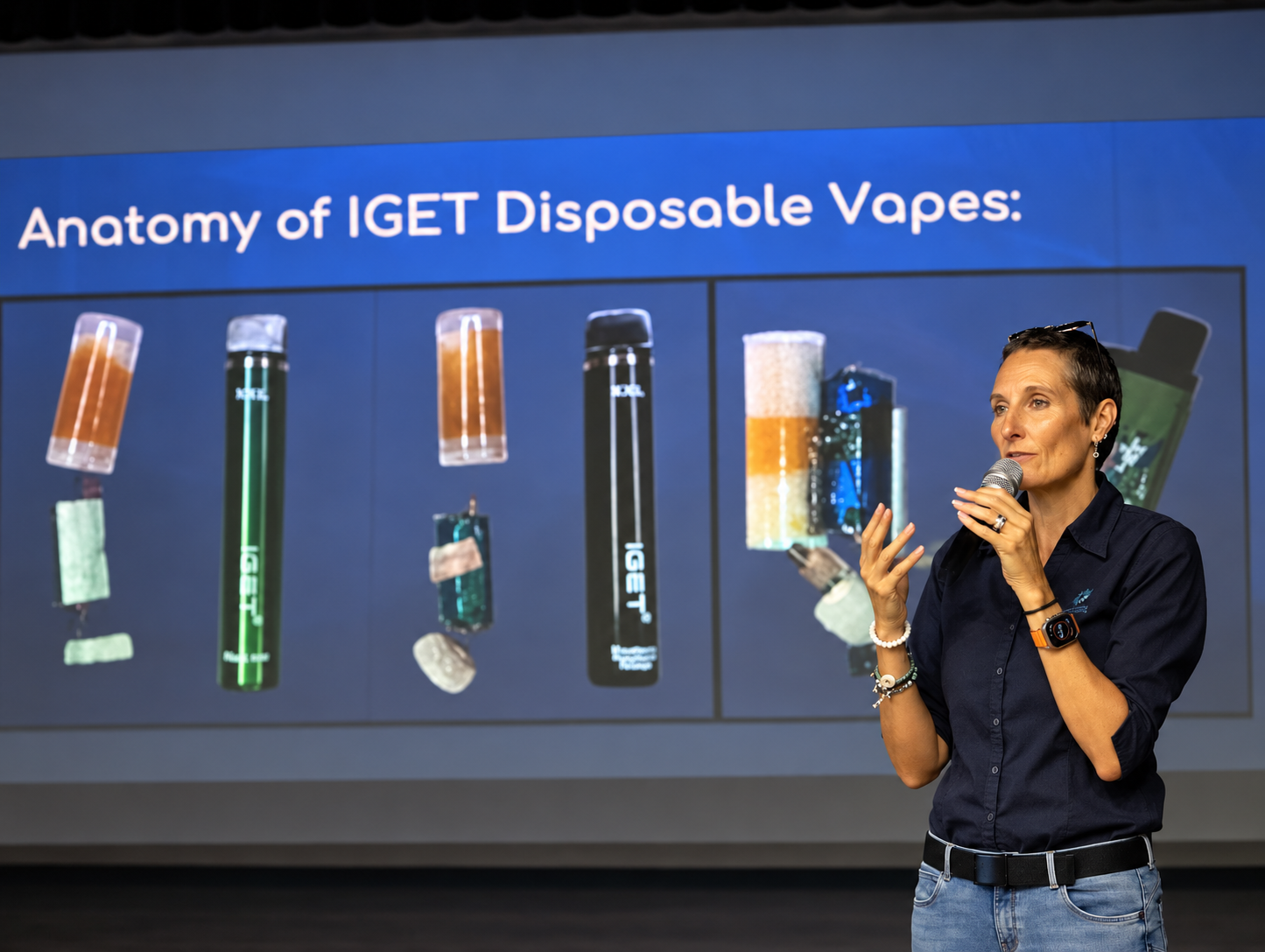 Azelene Williams with short dark hair, wearing a dark blue shirt, explaining the anatomy of IGET disposable vapes during a presentation. 