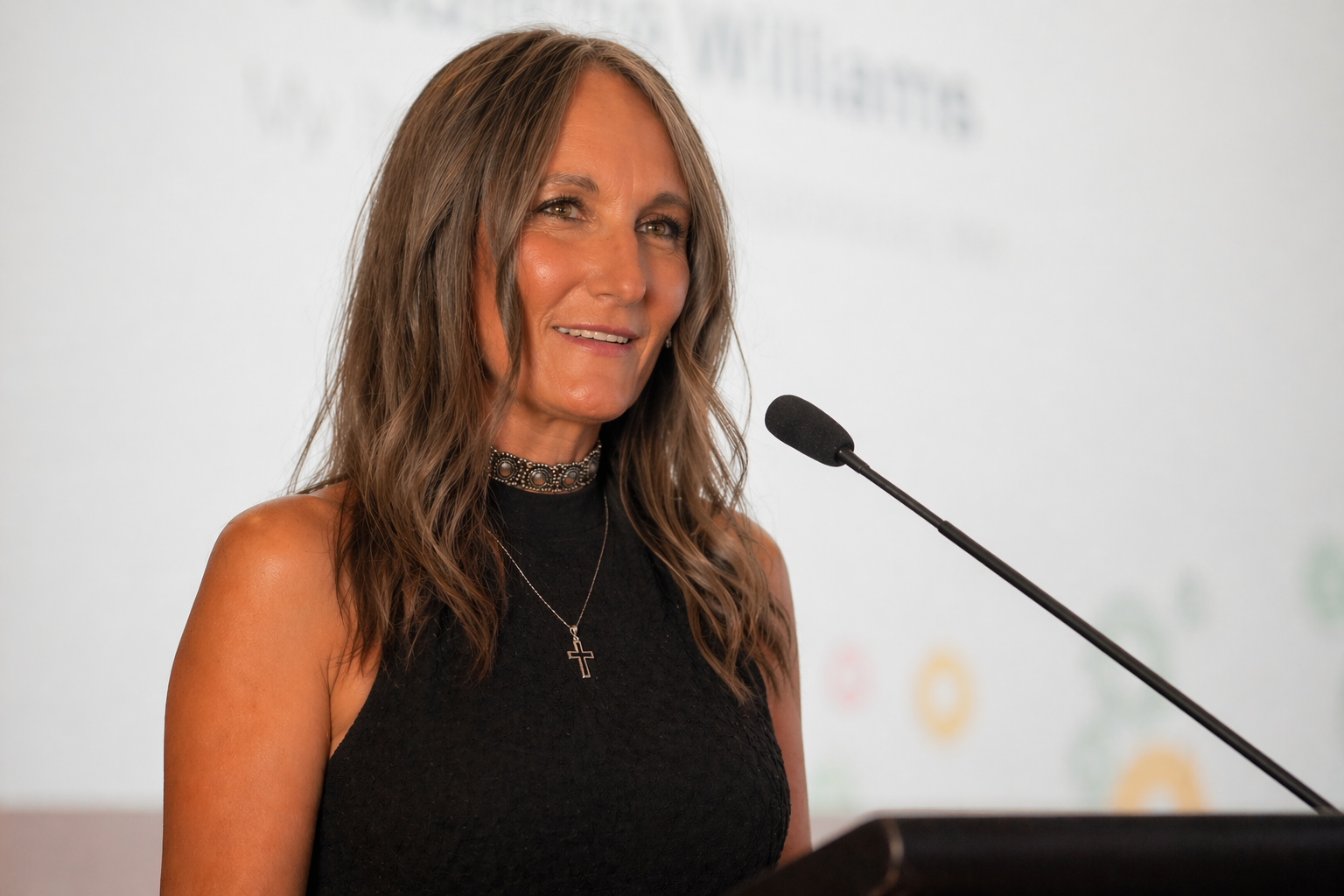Woman speaking at a podium with a microphone, wearing a black sleeveless top and a choker necklace with a cross pendant.